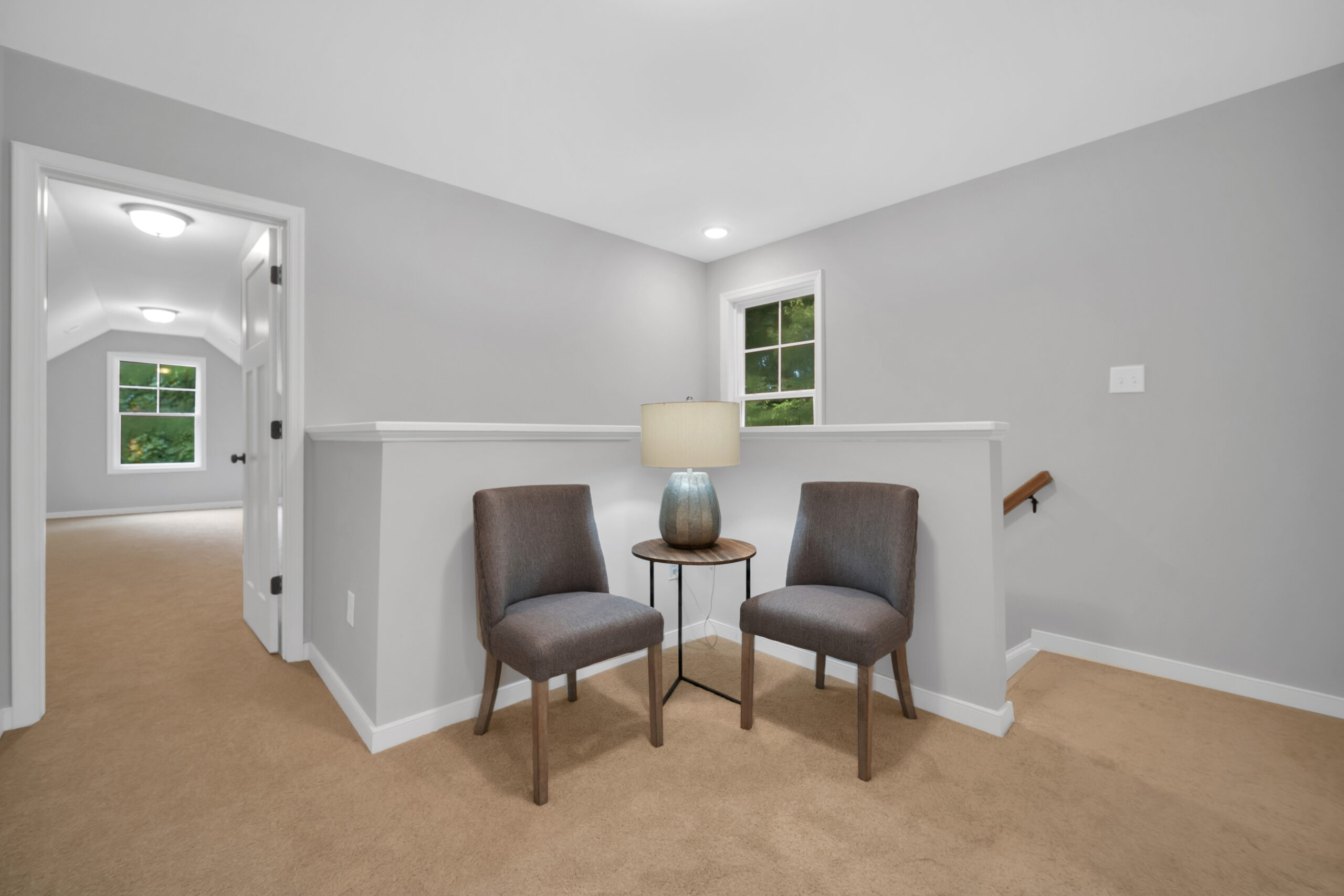 Interior of a modern home hallway featuring two chairs, a small table with a lamp, beige carpet, and white walls with two windows.