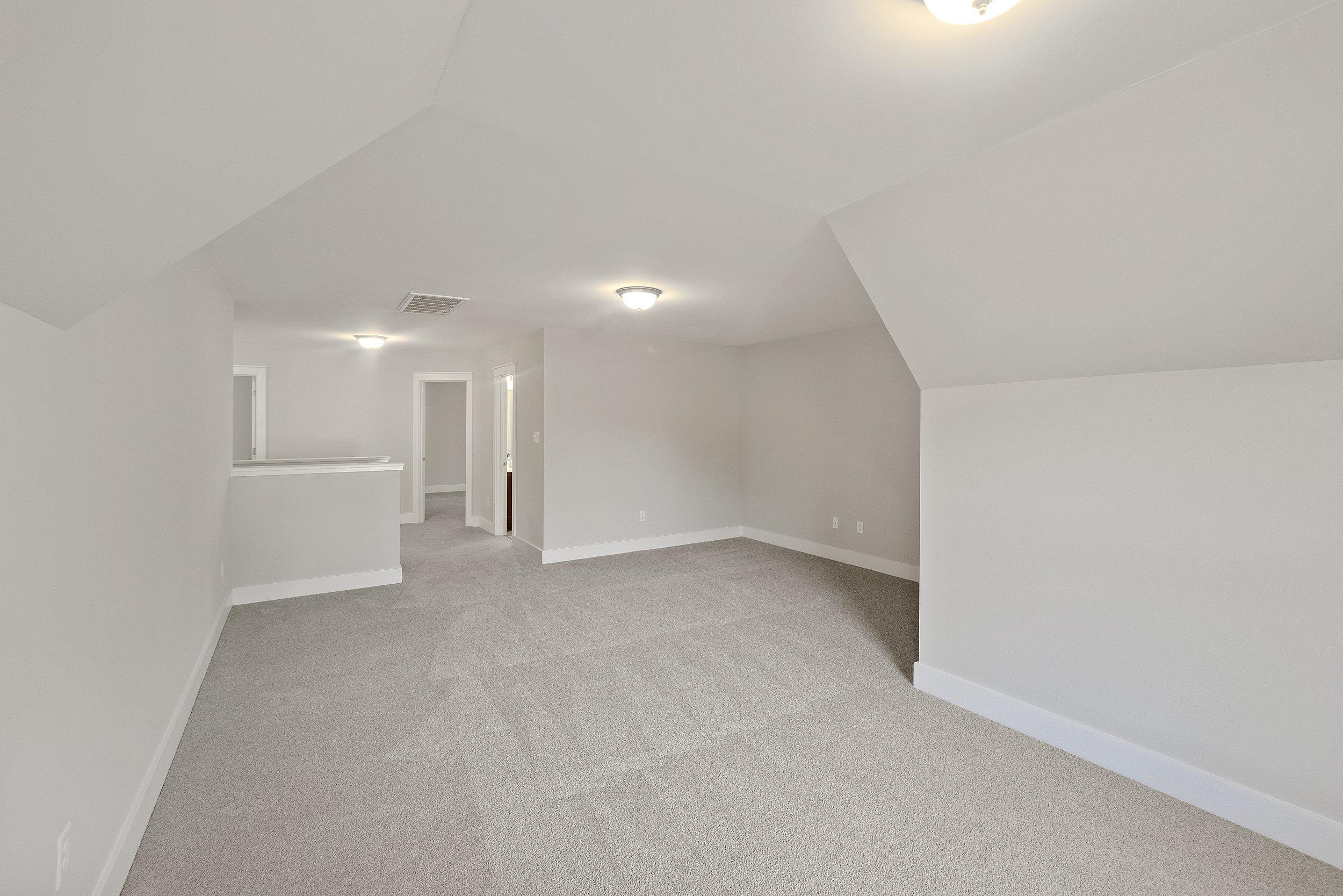 An empty room with beige carpet, white walls, and a slanted ceiling. There are two ceiling lights, a doorway, and a small partition wall in the background.