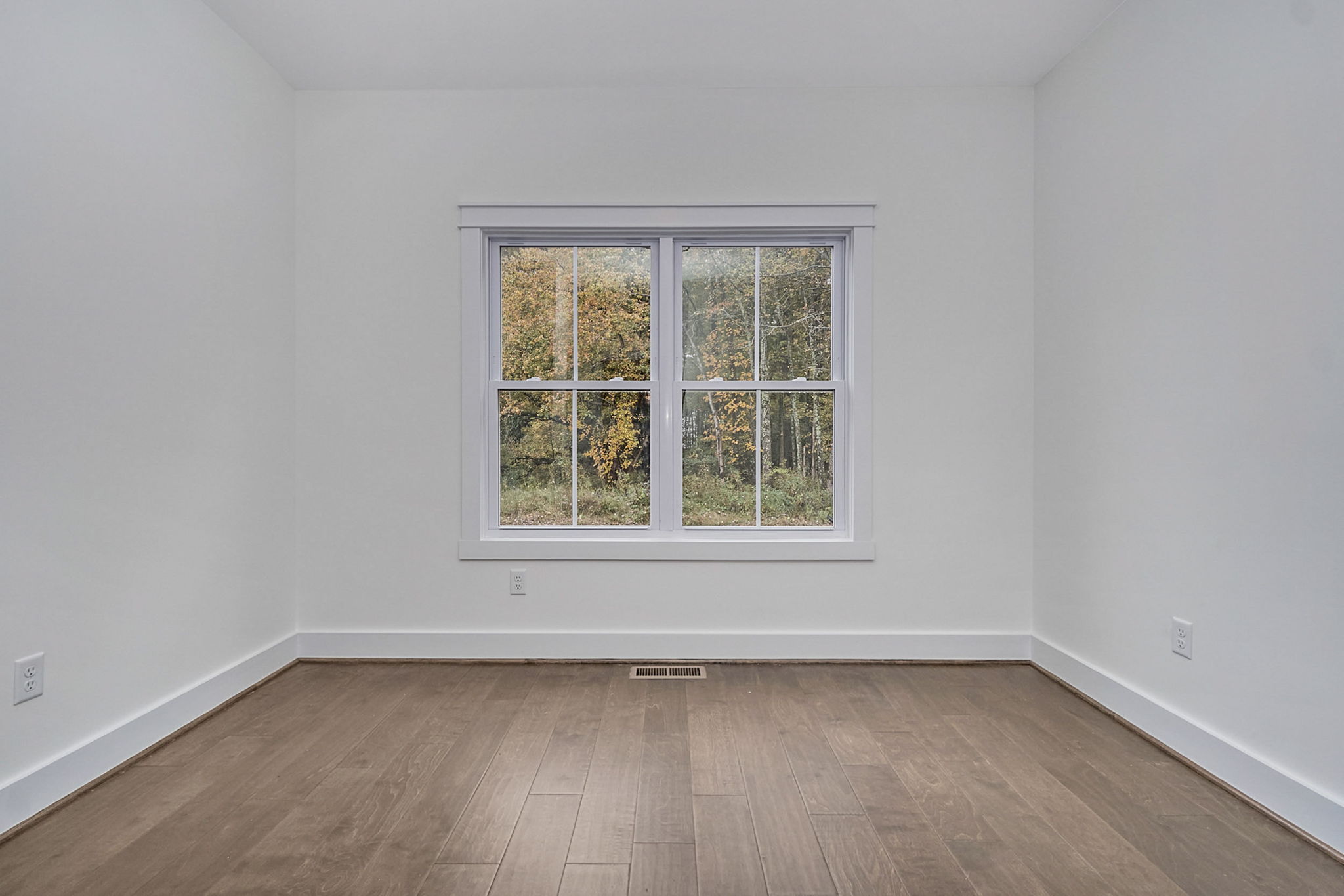 A small, empty room with white walls, hardwood floor, and a central window showing a view of trees outside.