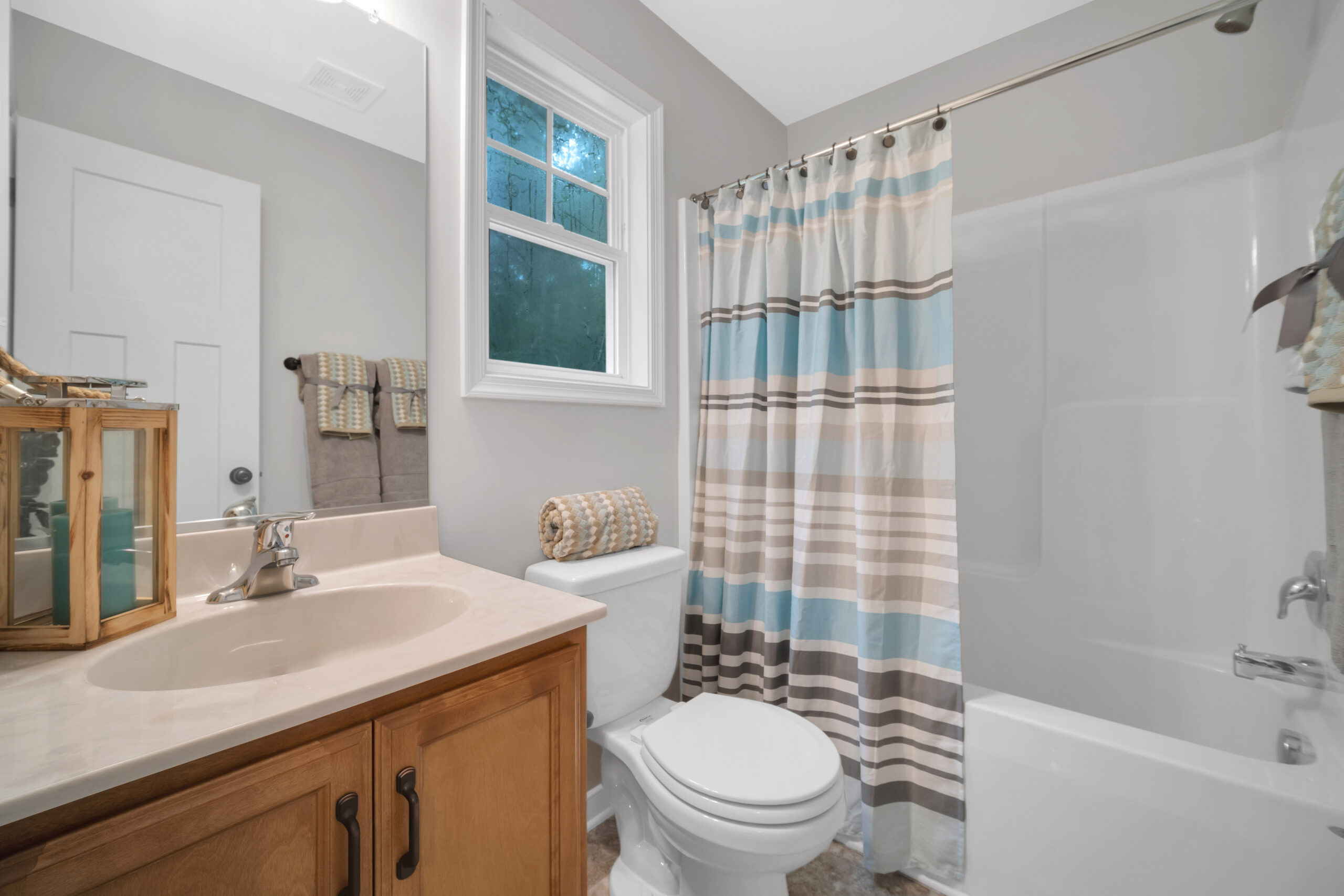 A small bathroom with a sink and wooden cabinet on the left, a toilet in the center, and a bathtub with a striped shower curtain on the right. There is a window above the toilet.