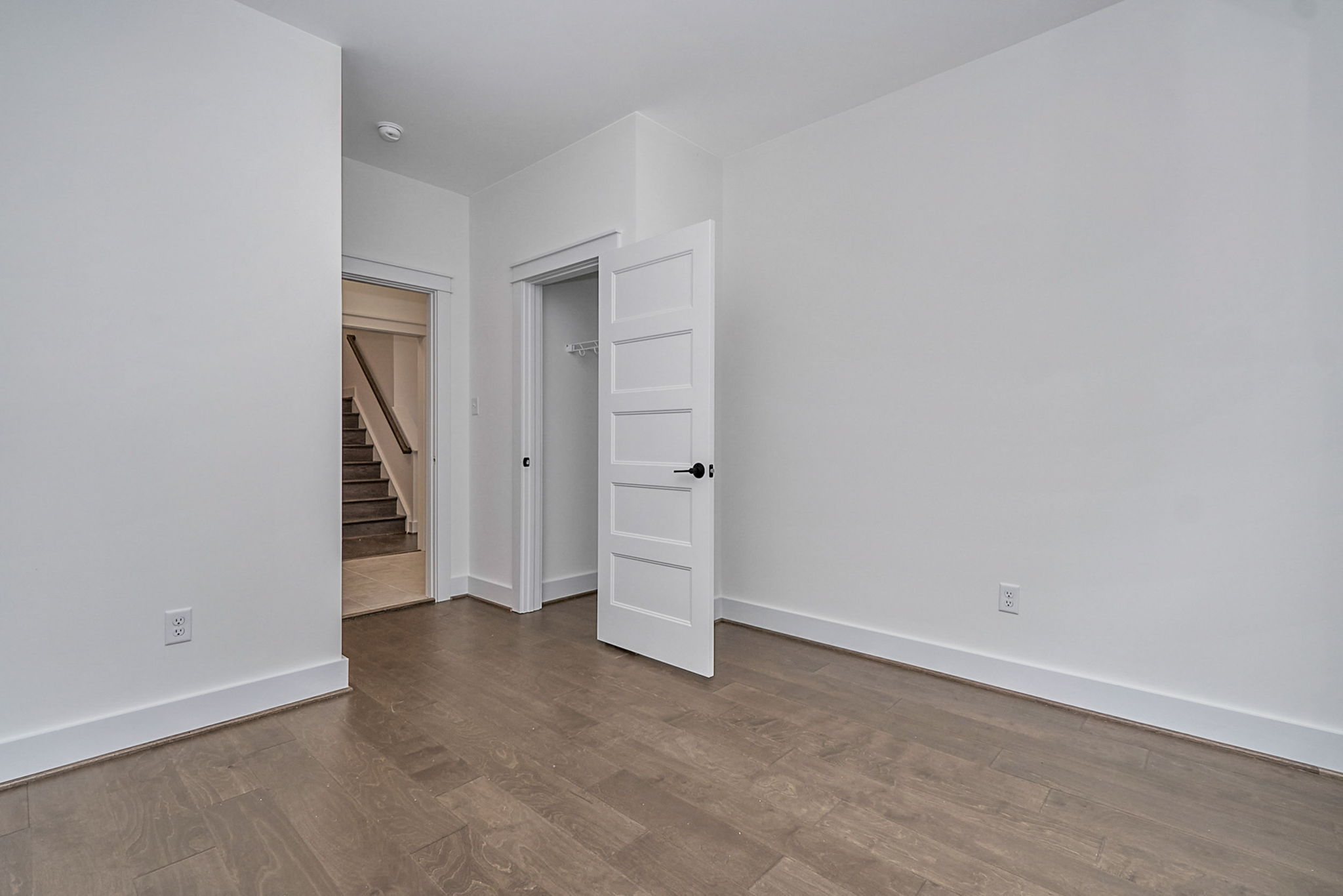 Empty room with light gray walls, wooden flooring, and a white door slightly ajar. An open doorway leads to a staircase. A dark handle is visible on the door, and electrical outlets are on the walls.