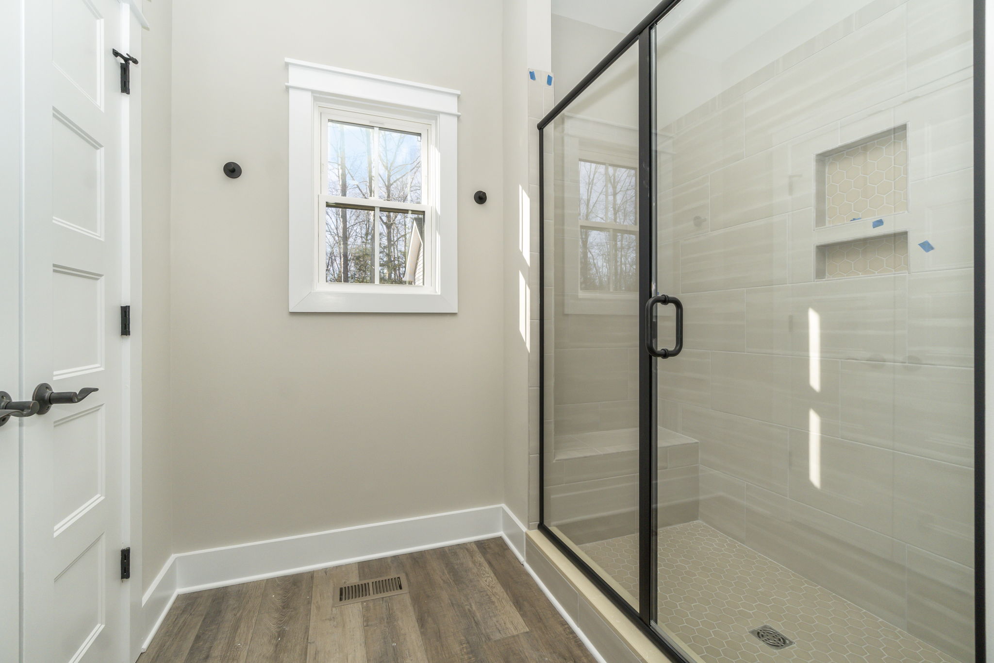 A modern bathroom featuring a glass shower enclosure, beige tiles, a small window, and wooden flooring.