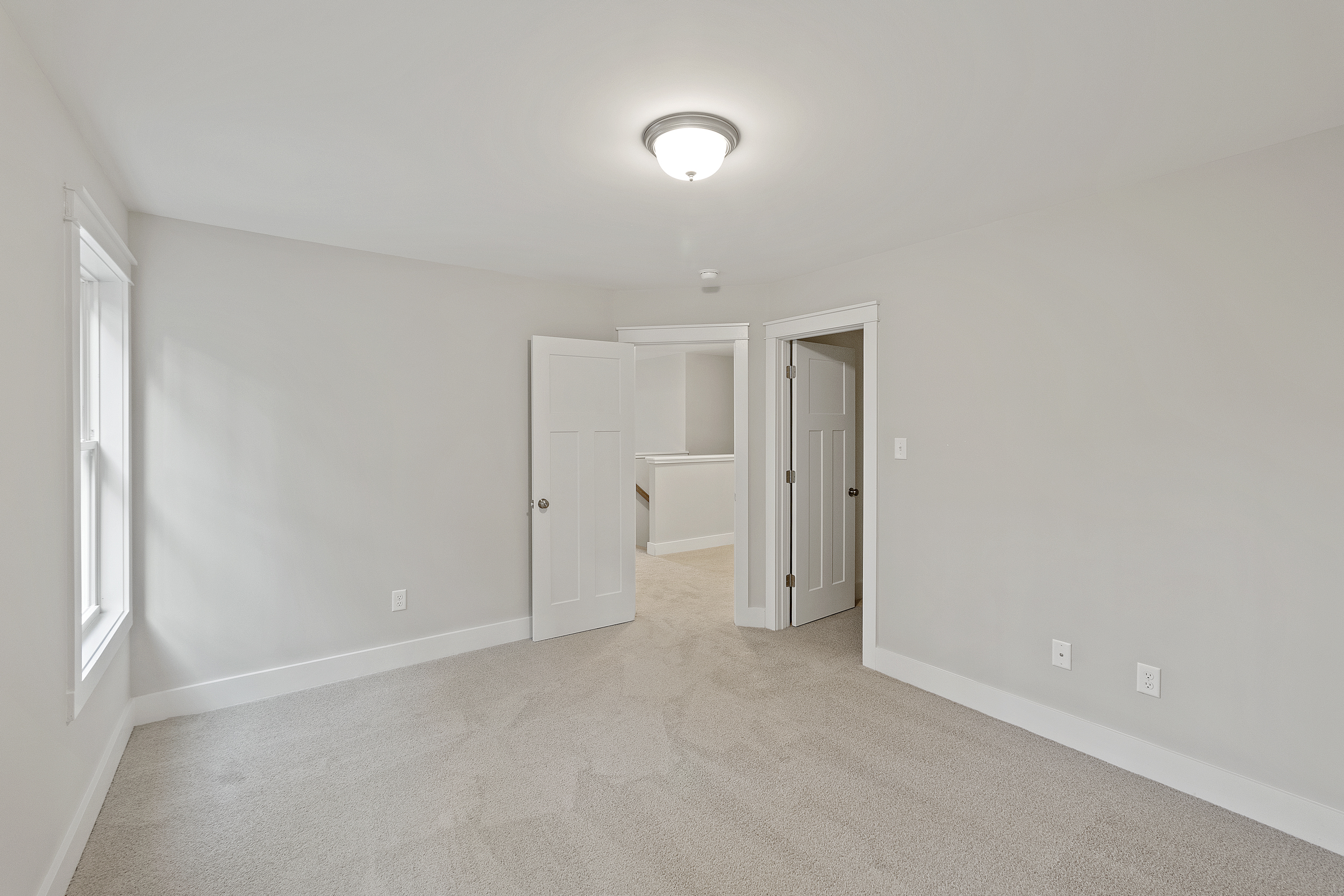Empty room with beige carpet, white walls, and a ceiling light fixture. The room has a window on the left, a partially open door leading to a hallway, and electrical outlets on the right wall.