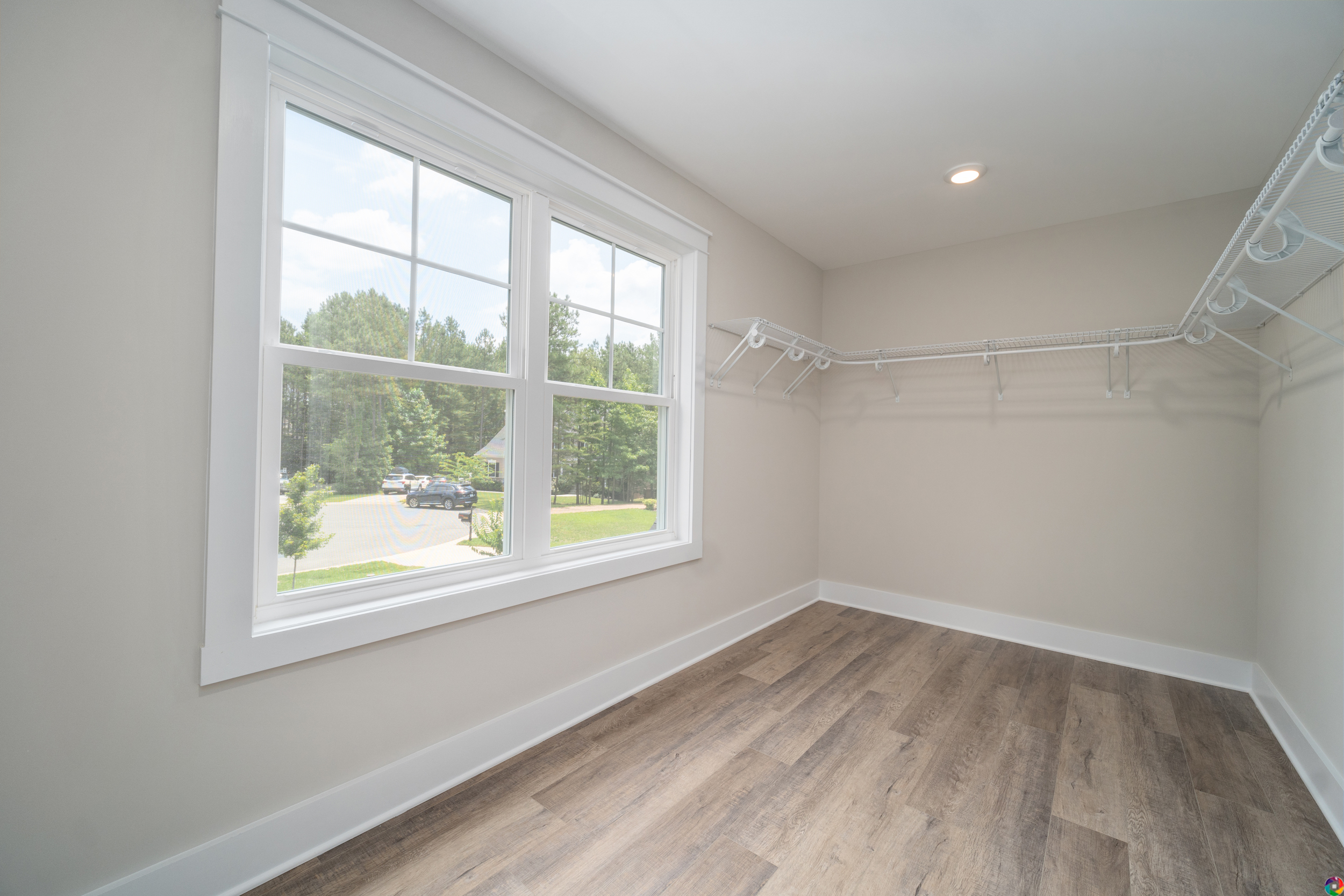A walk-in closet with light wood flooring, beige walls, multiple white wire shelves, and a large window with a view of trees and a driveway.