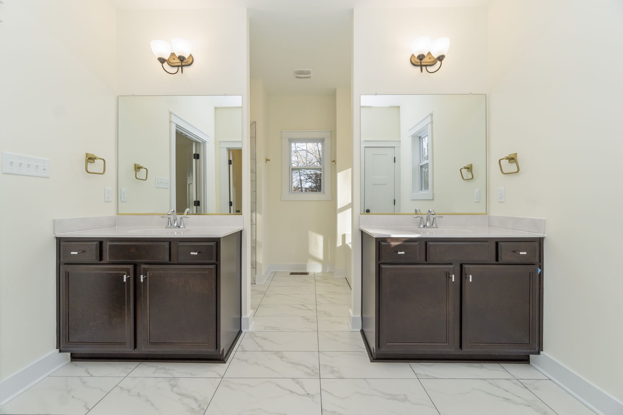 A bright, symmetrical bathroom with two dark wooden vanities, marble floors, mirrors, and wall-mounted lights.