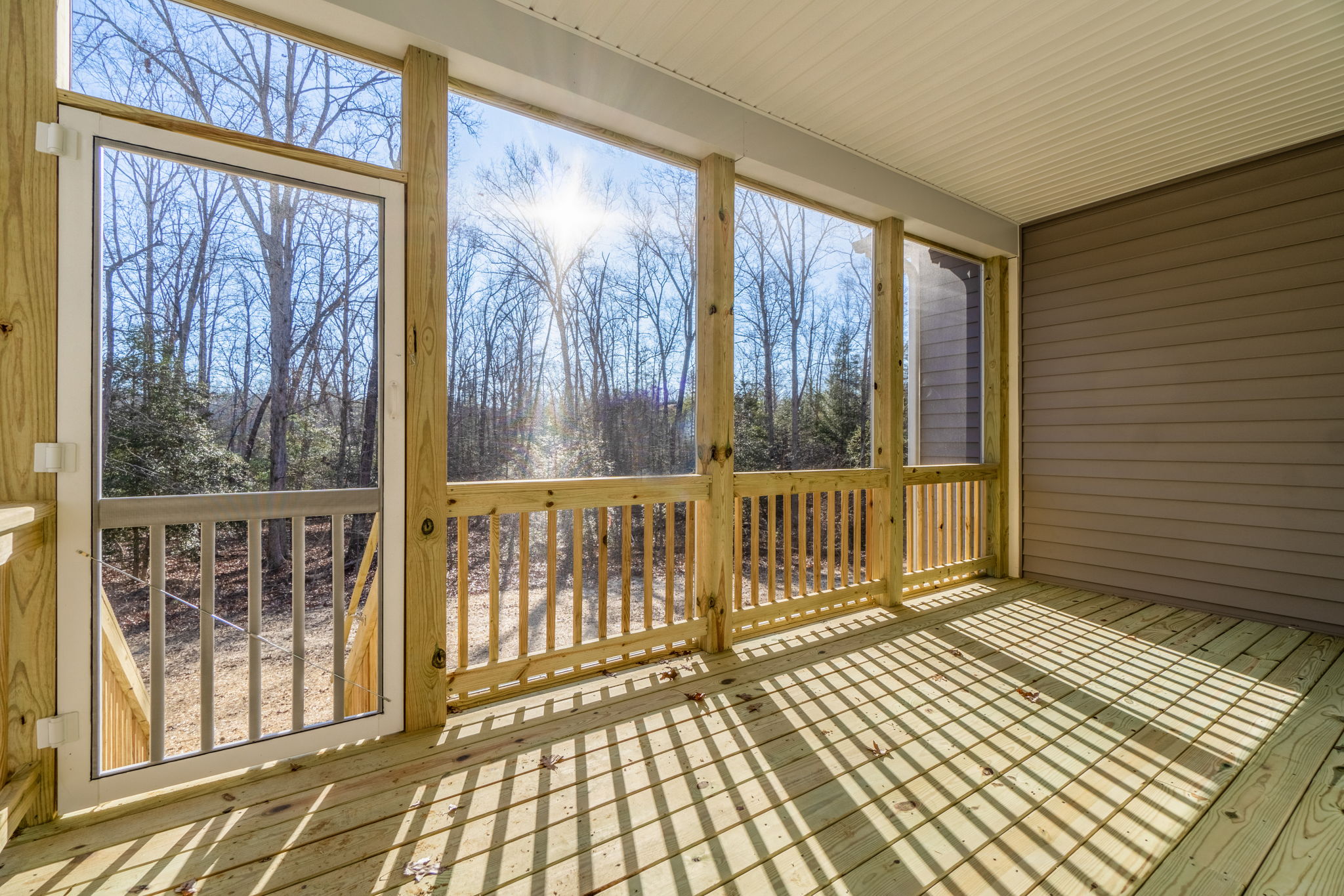 Bright sunlight streams into a spacious screened porch with wooden floors and railings, showcasing the surrounding leafless trees.