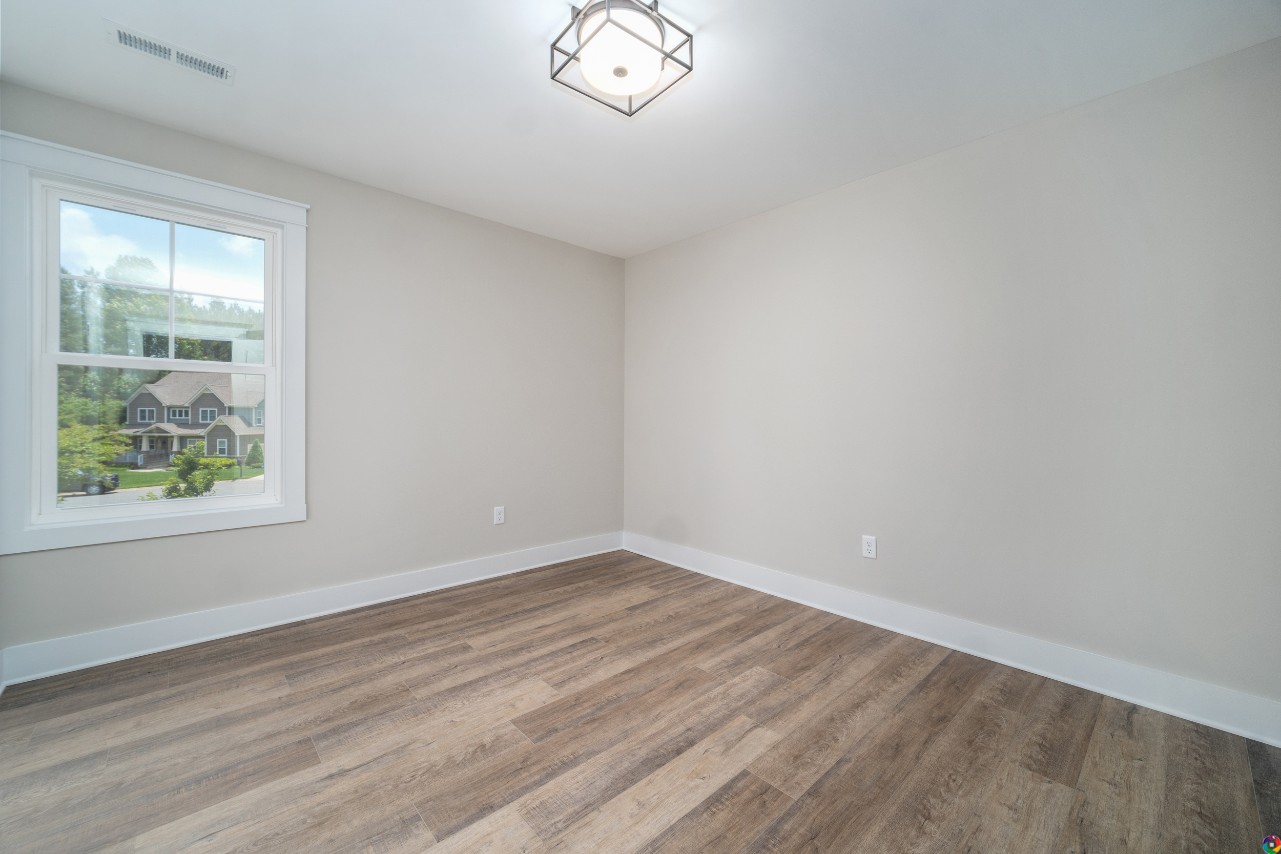A vacant room with light-colored walls, a large window showing a view of houses and greenery, hardwood floors, and a modern ceiling light fixture.