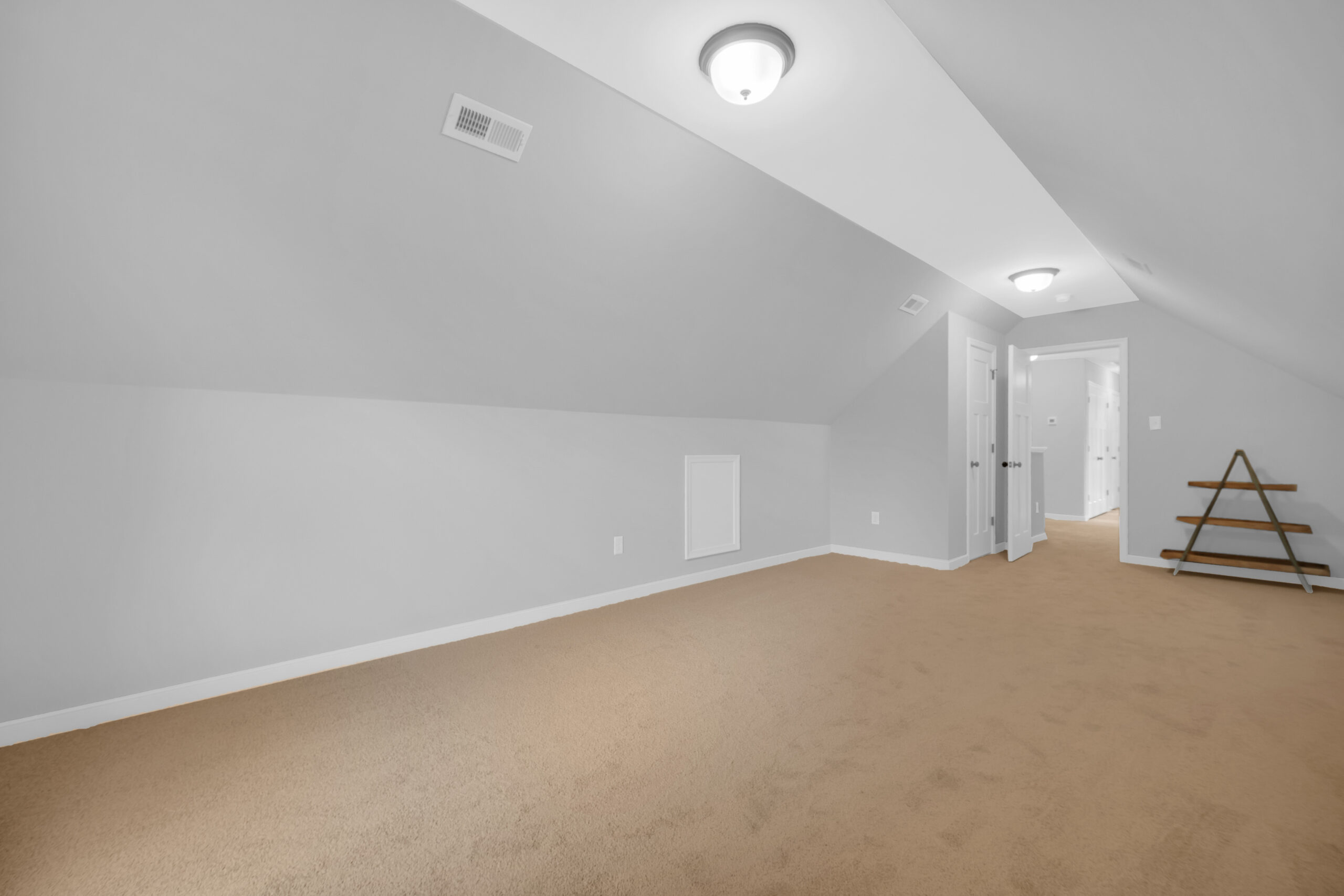 Spacious, well-lit attic room with beige carpeting, slanted white walls, recessed lighting, and an open door at the end.