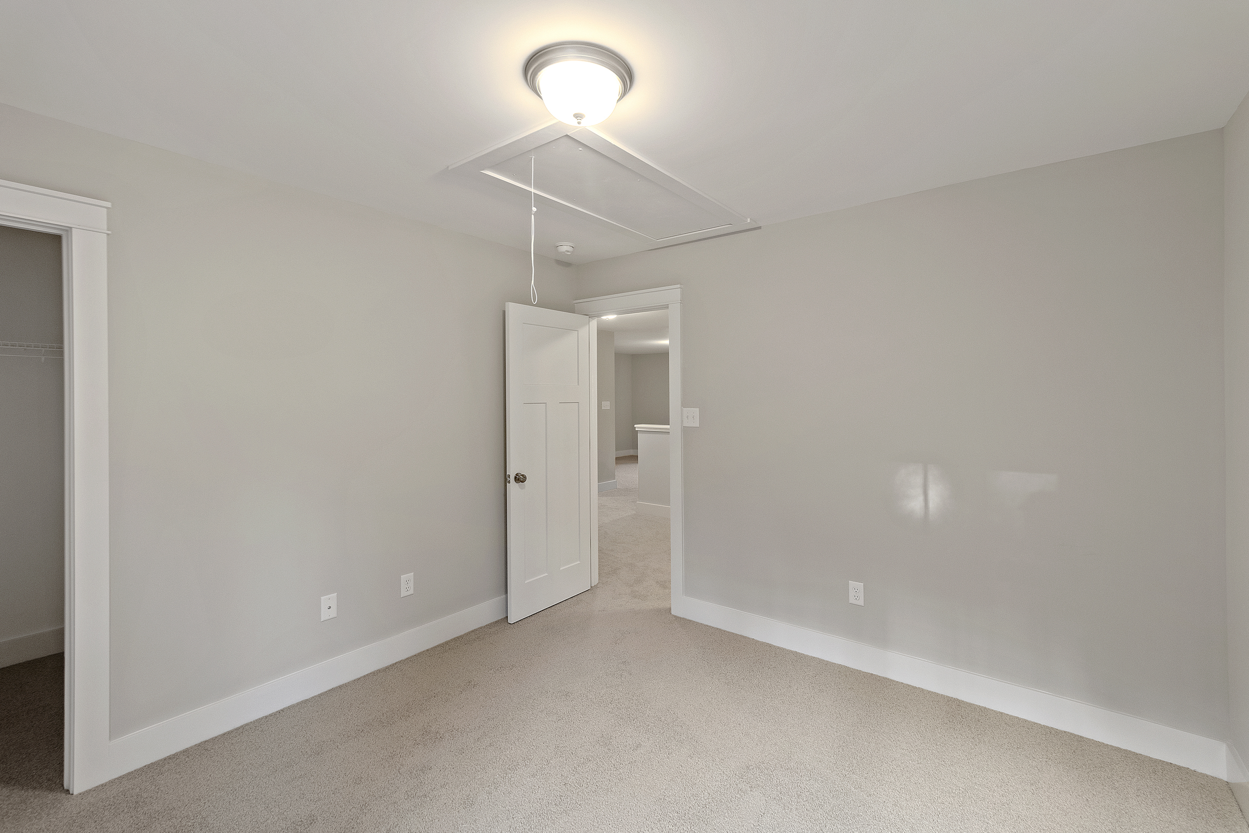 Empty room with beige carpet, light gray walls, a closed white door, a closet, a ceiling light fixture, and a visible staircase in the adjoining room.