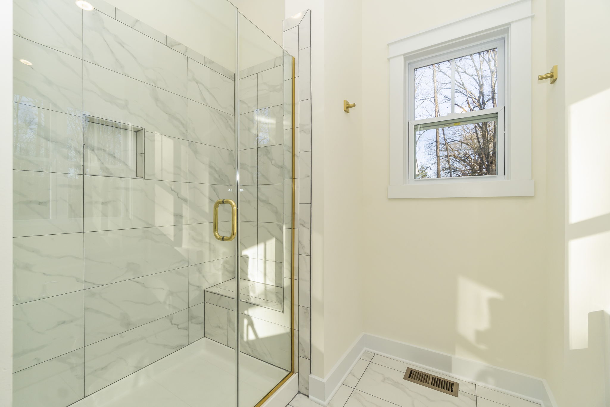 A modern bathroom featuring marble tiles with a glass shower enclosure, gold fixtures, and a window allowing natural light.