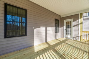 Wooden porch of a house with white siding, featuring a door and two windows, casting shadows in bright sunlight.