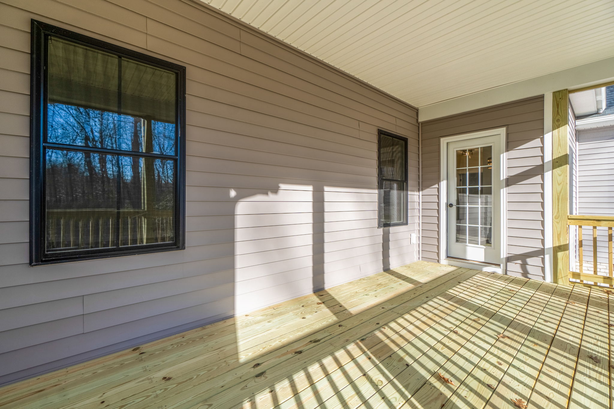 Wooden porch of a house with white siding, featuring a door and two windows, casting shadows in bright sunlight.