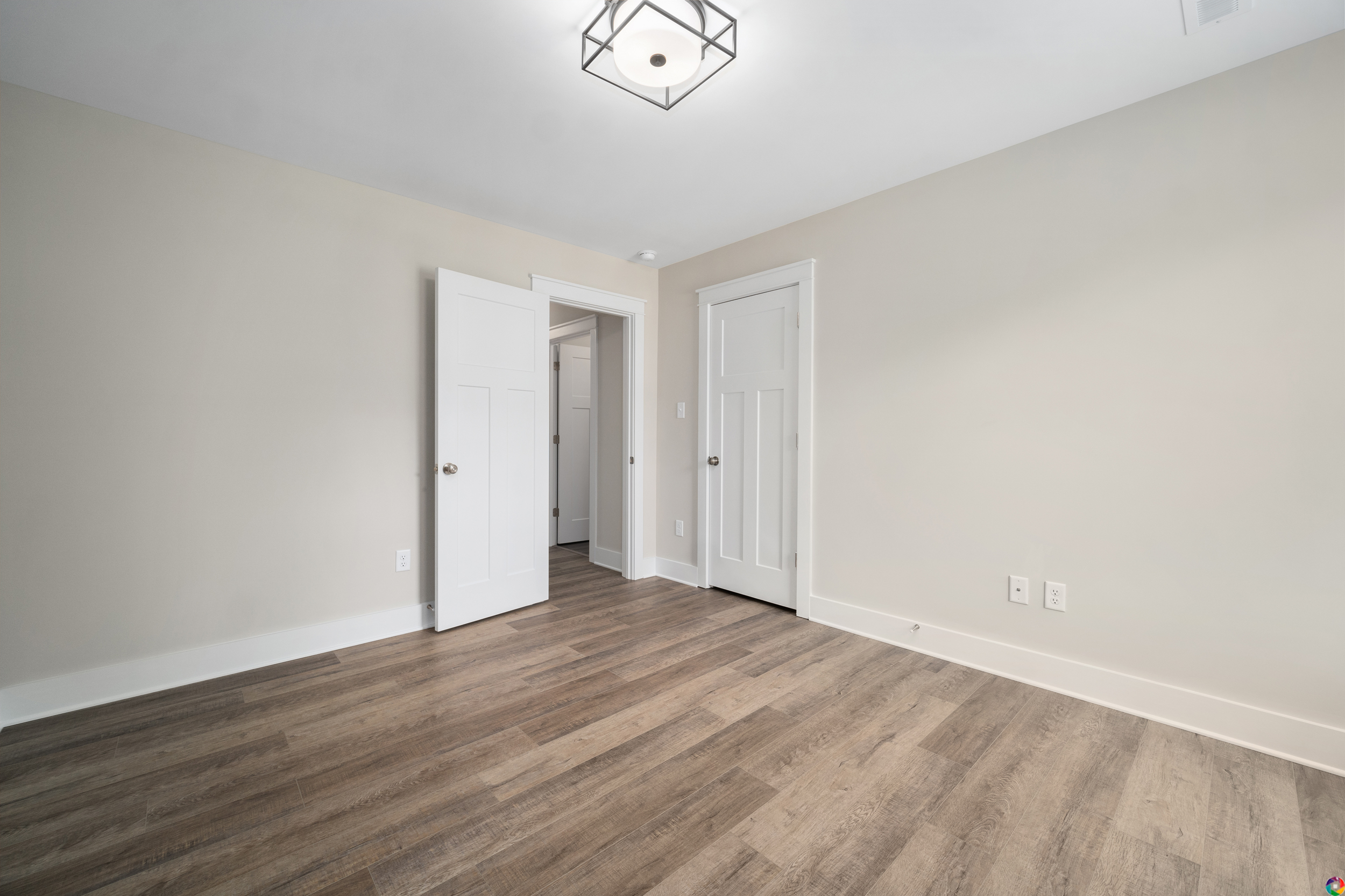 A vacant room with light-colored walls, wooden flooring, and two white doors; one door is open, showing a small closet. A ceiling light fixture is visible.