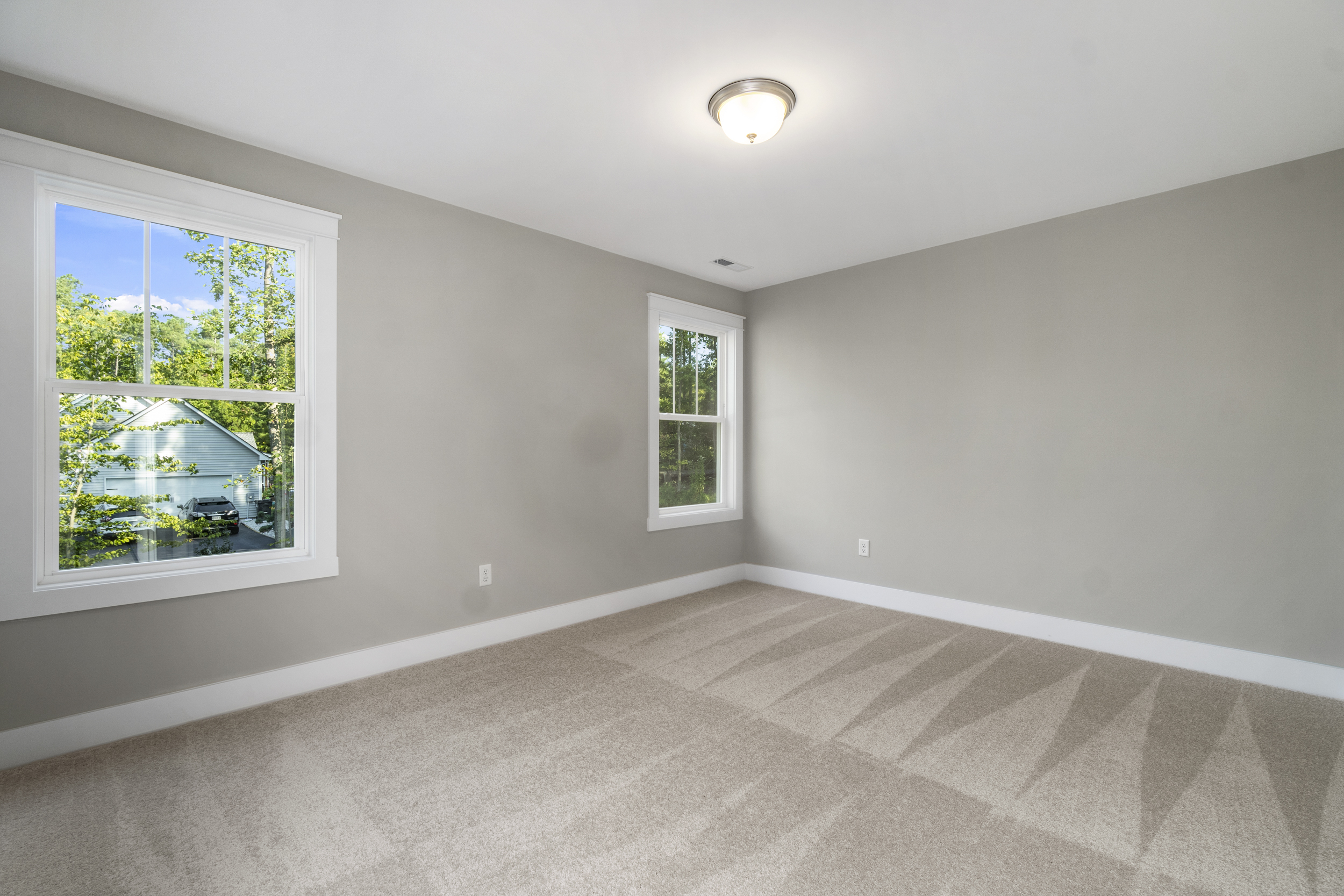 A bright, empty room with beige carpet, gray walls, and two windows that allow natural light to fill the space. A ceiling light fixture is centered above. The view through the windows shows green trees.