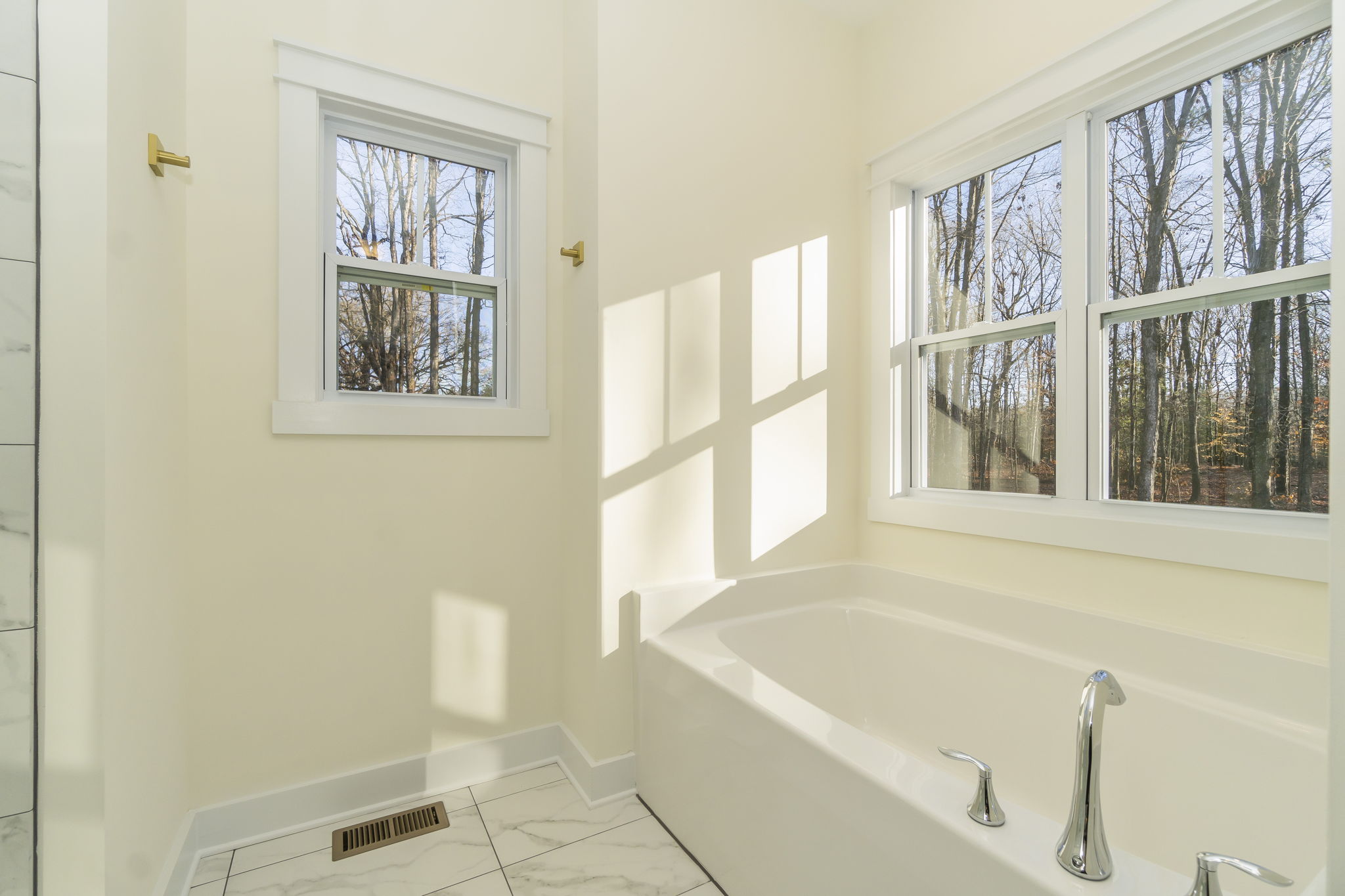 A bright bathroom with a soaking tub under large windows showing a view of leafless trees, white walls, and a tiled floor.