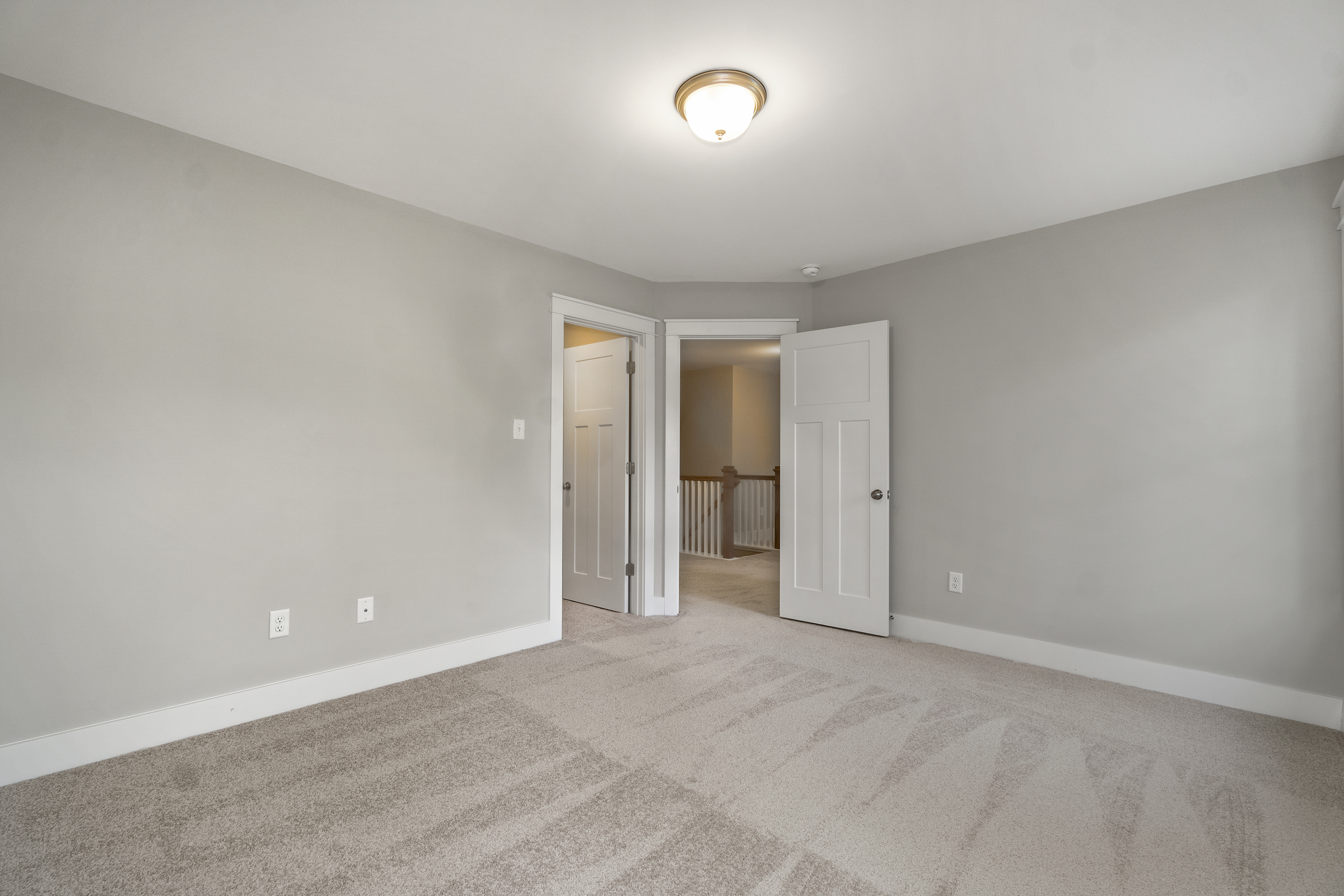 A clean and empty carpeted room with beige walls, white trim, a light fixture on the ceiling, and an open door leading to a hallway with a staircase. Two electrical outlets are visible on the wall.