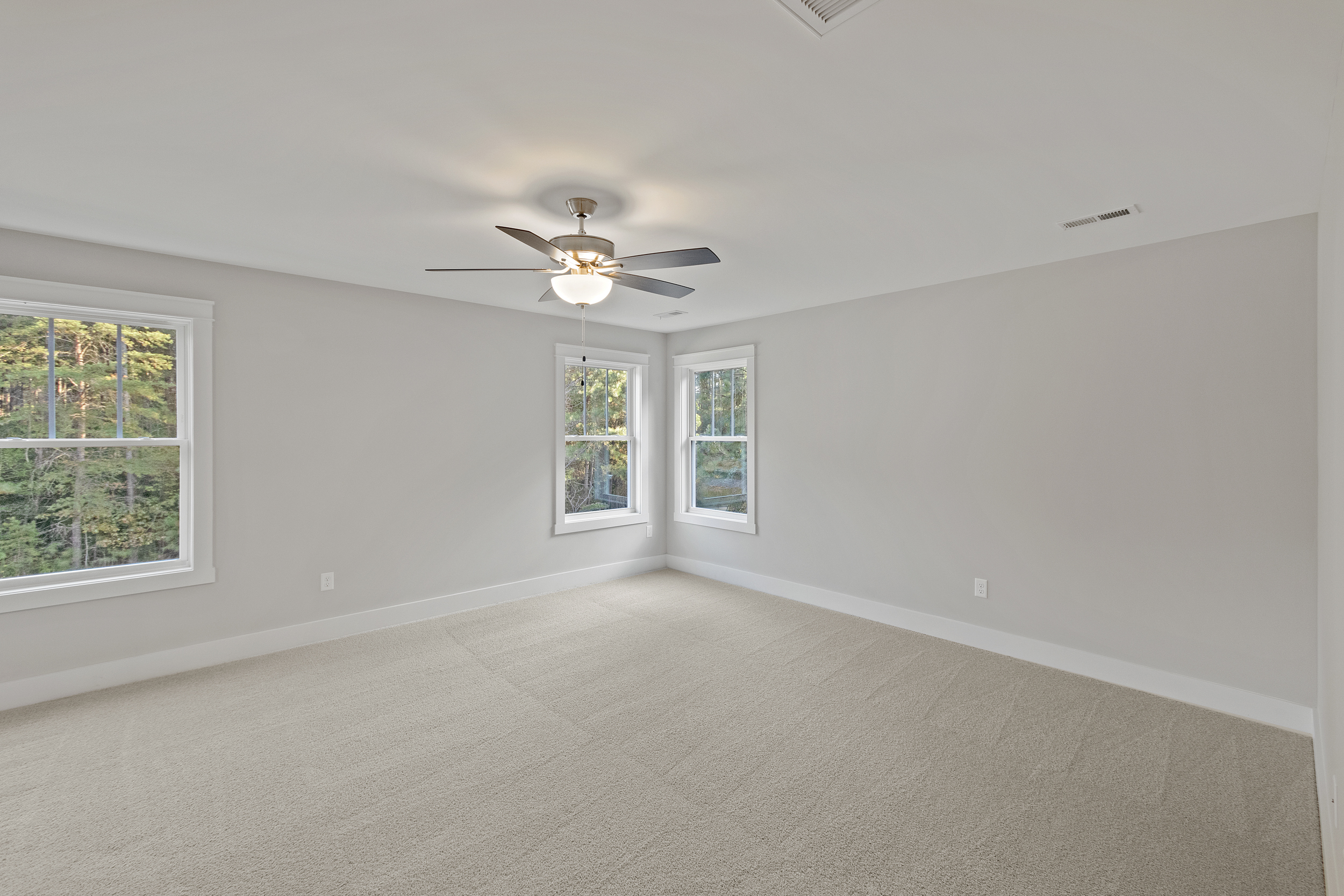 A spacious, empty room with beige carpet, white walls, a ceiling fan with lights, and two double windows letting in natural light.