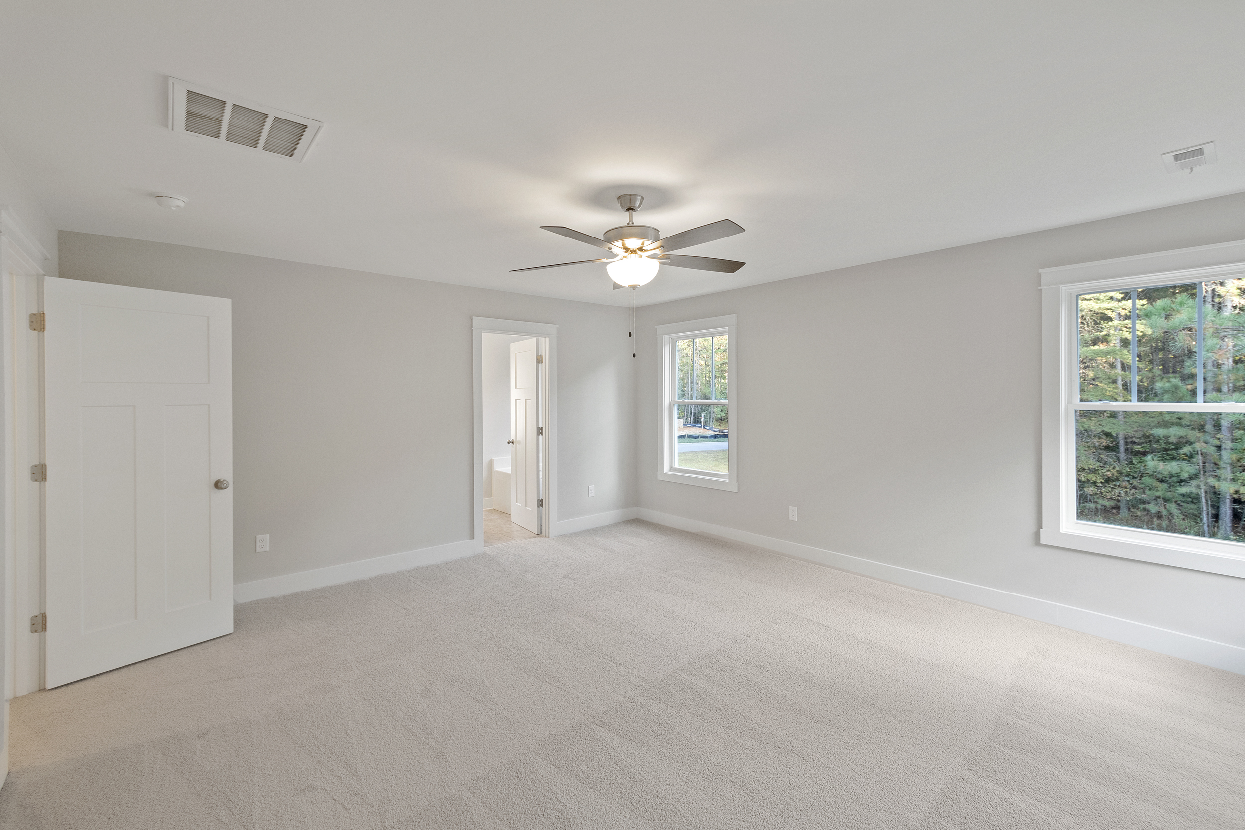 Spacious empty bedroom with light gray walls, a white ceiling fan, and two large windows. There's a door leading to an adjoining bathroom. The room is carpeted in a light beige color.