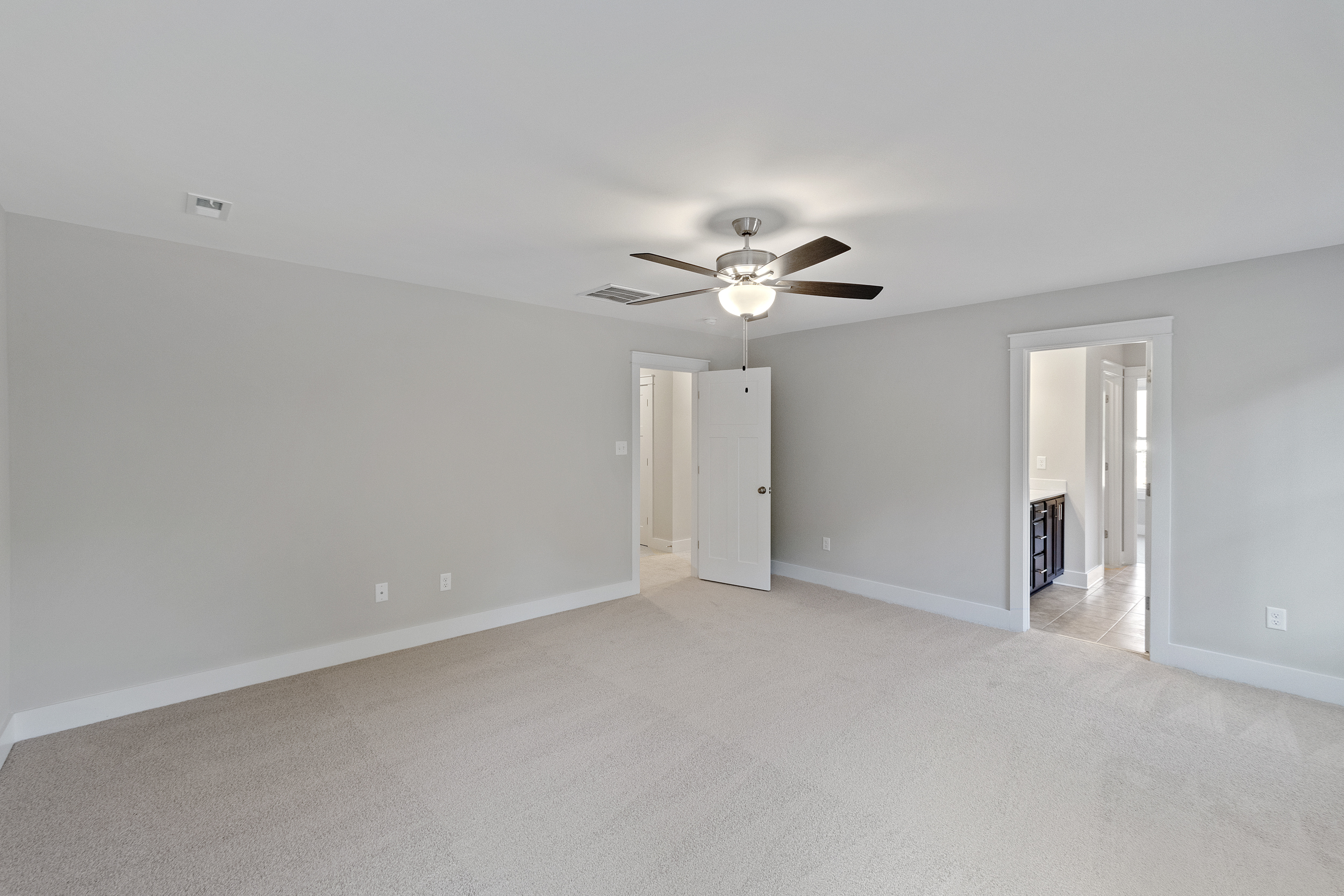 A spacious, empty room with light-colored carpet, gray walls, a ceiling fan with light, a white door slightly ajar, and an open doorway leading to another room with tiled flooring.