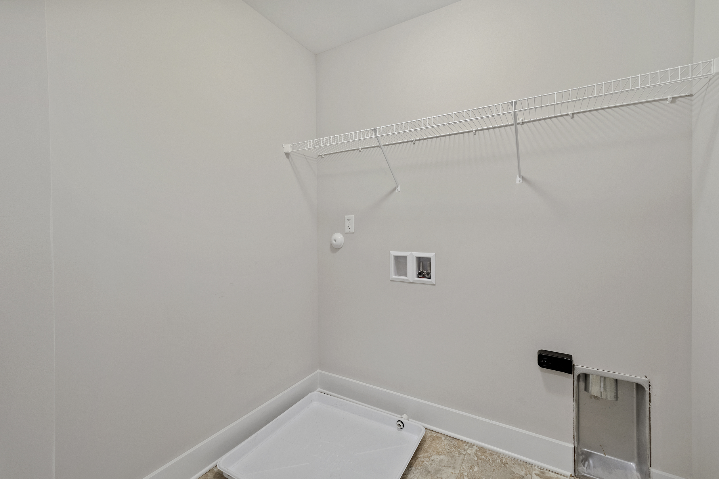 Empty laundry room with white walls, a utility sink basin, a wire rack shelf above, and outlets for washer and dryer connections.