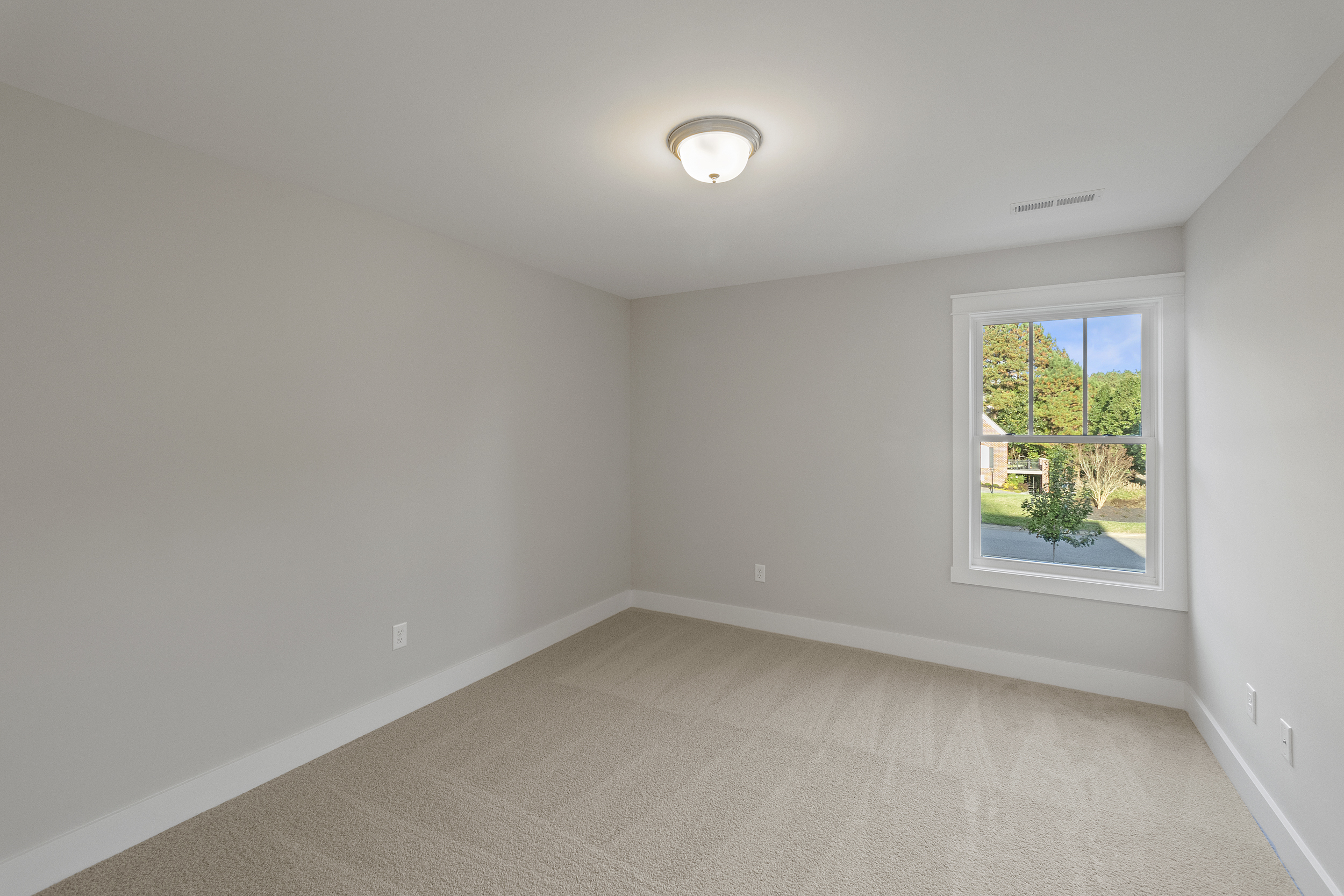 An empty, carpeted room with beige walls, a ceiling light fixture, and a window showing a view of greenery outside.