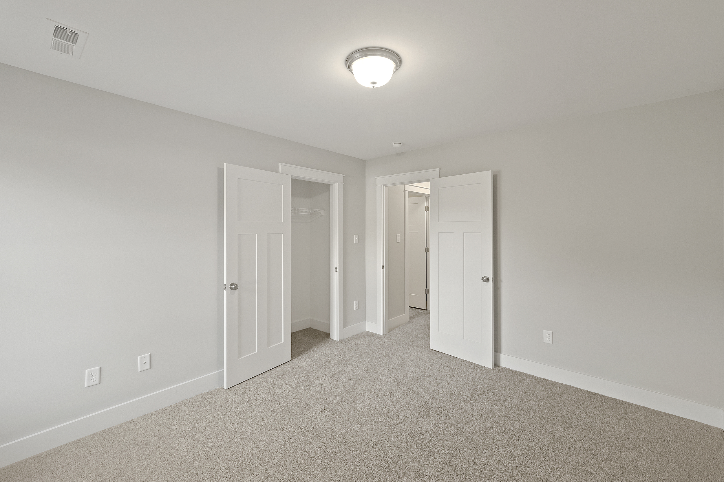 A clean, empty bedroom with carpeted floor, white walls, and a ceiling light. There are two open doors revealing closets and an adjacent room.