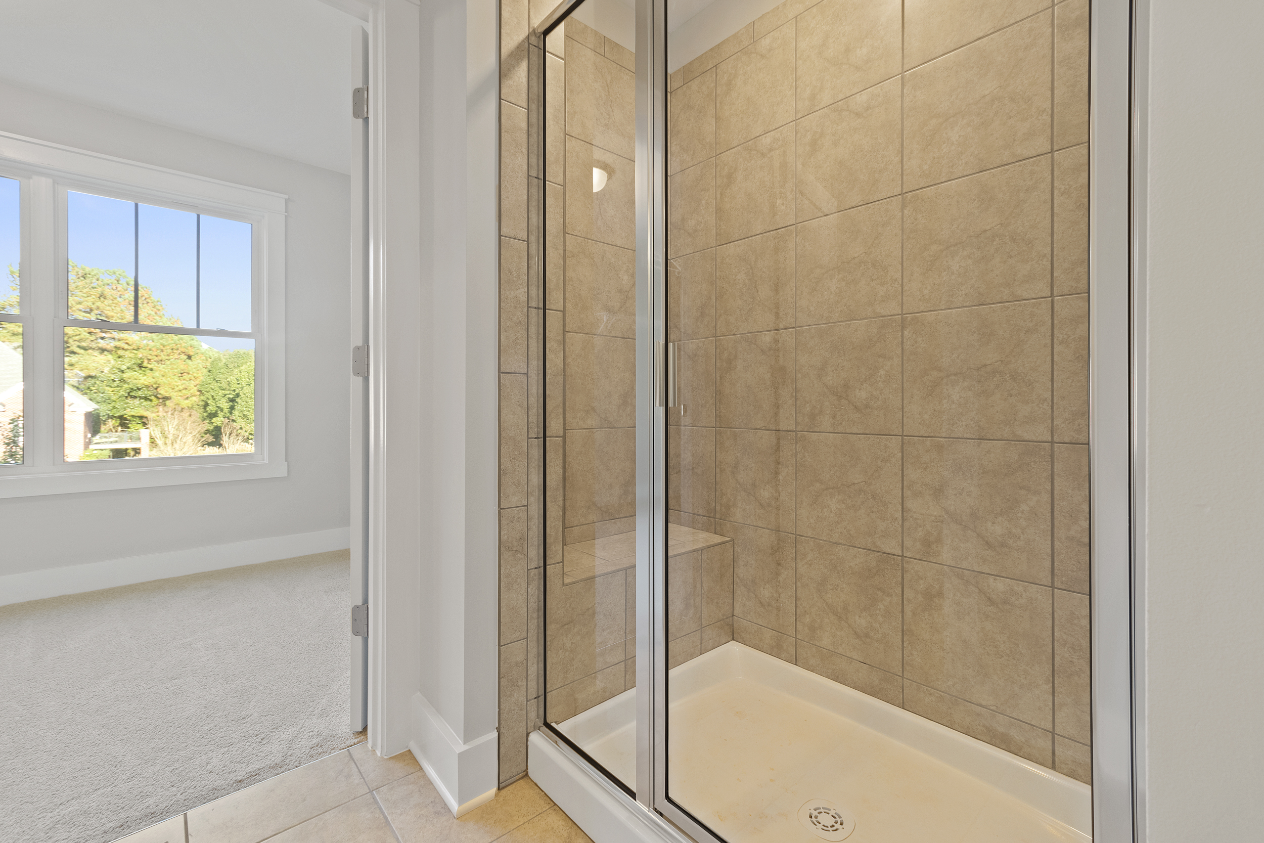 A bathroom with a glass-enclosed shower featuring beige tiles, adjacent to a room with a large window overlooking the outdoors.