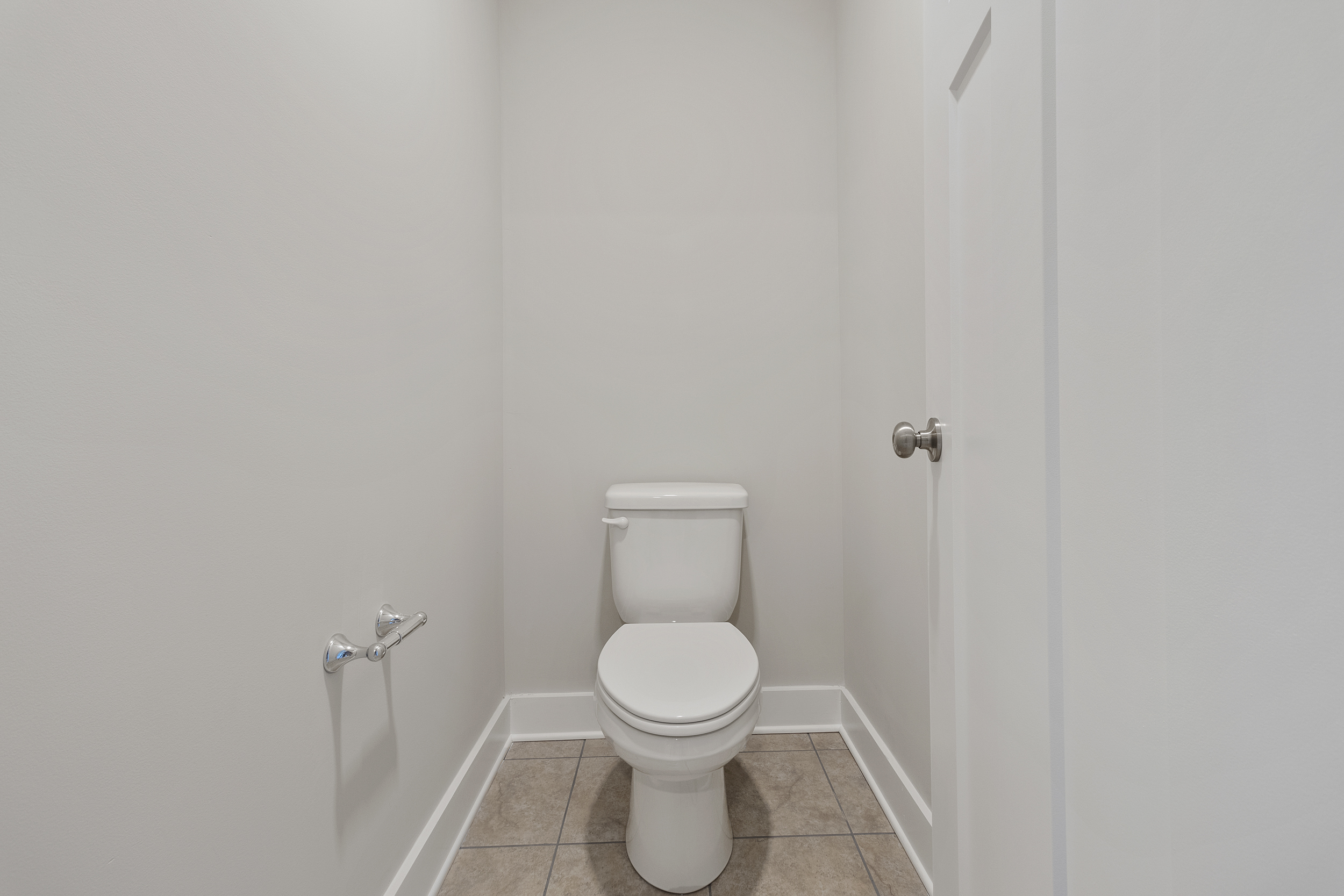 A small bathroom with a white toilet, empty toilet paper holder, gray tiled floor, and white walls.