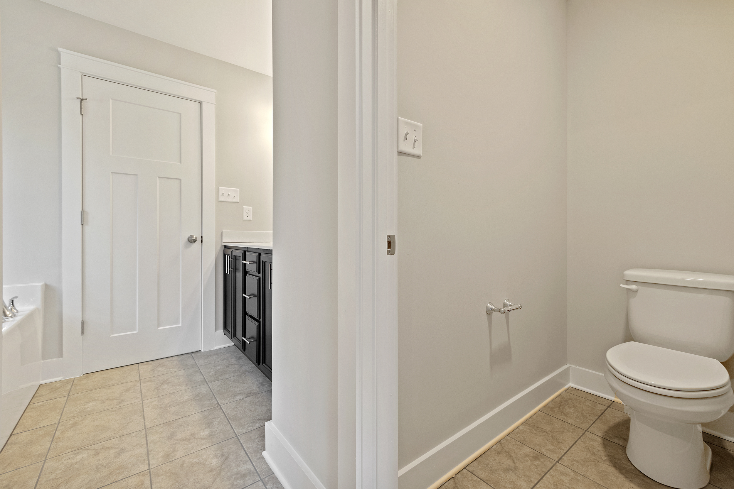 A bathroom with a toilet, a black vanity with a sink, a white door, beige tiled floor, and light gray walls.