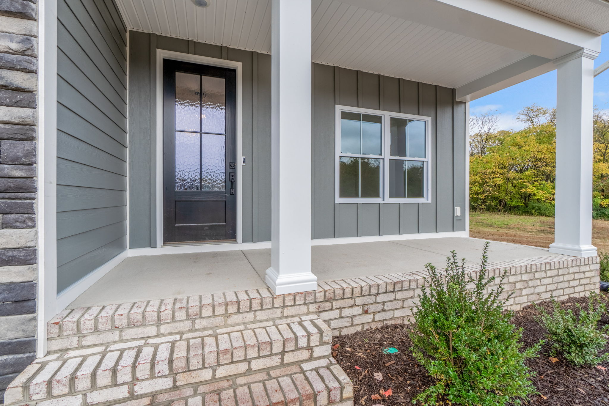 Front porch of a house featuring a black front door with frosted glass, white columns, gray siding, and a brick staircase with a small landscaped area containing a few shrubs.