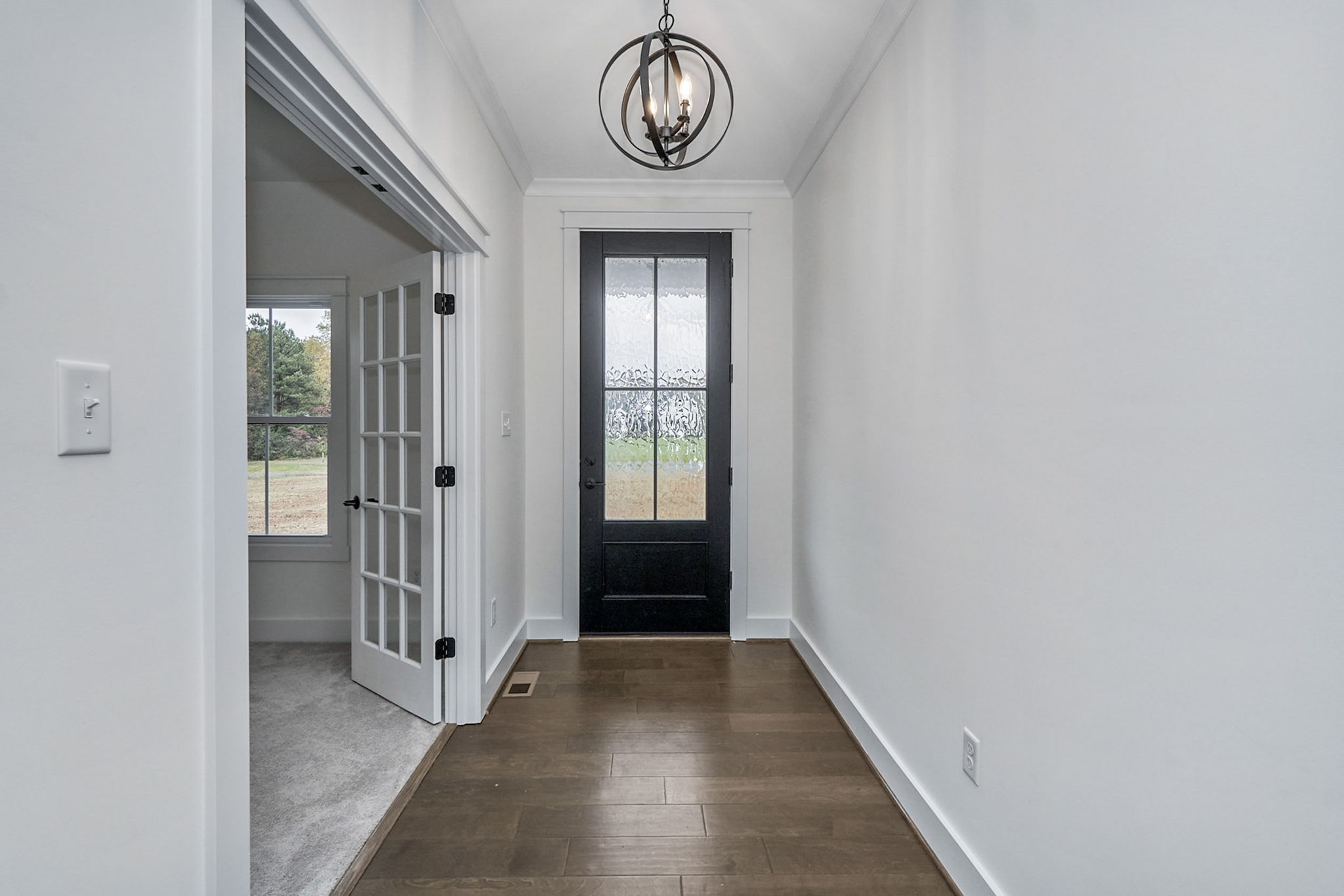 A narrow hallway with hardwood floors features a black front door with glass panes. To the left, there is a room with French doors and a carpeted floor. A modern hanging light fixture is overhead.