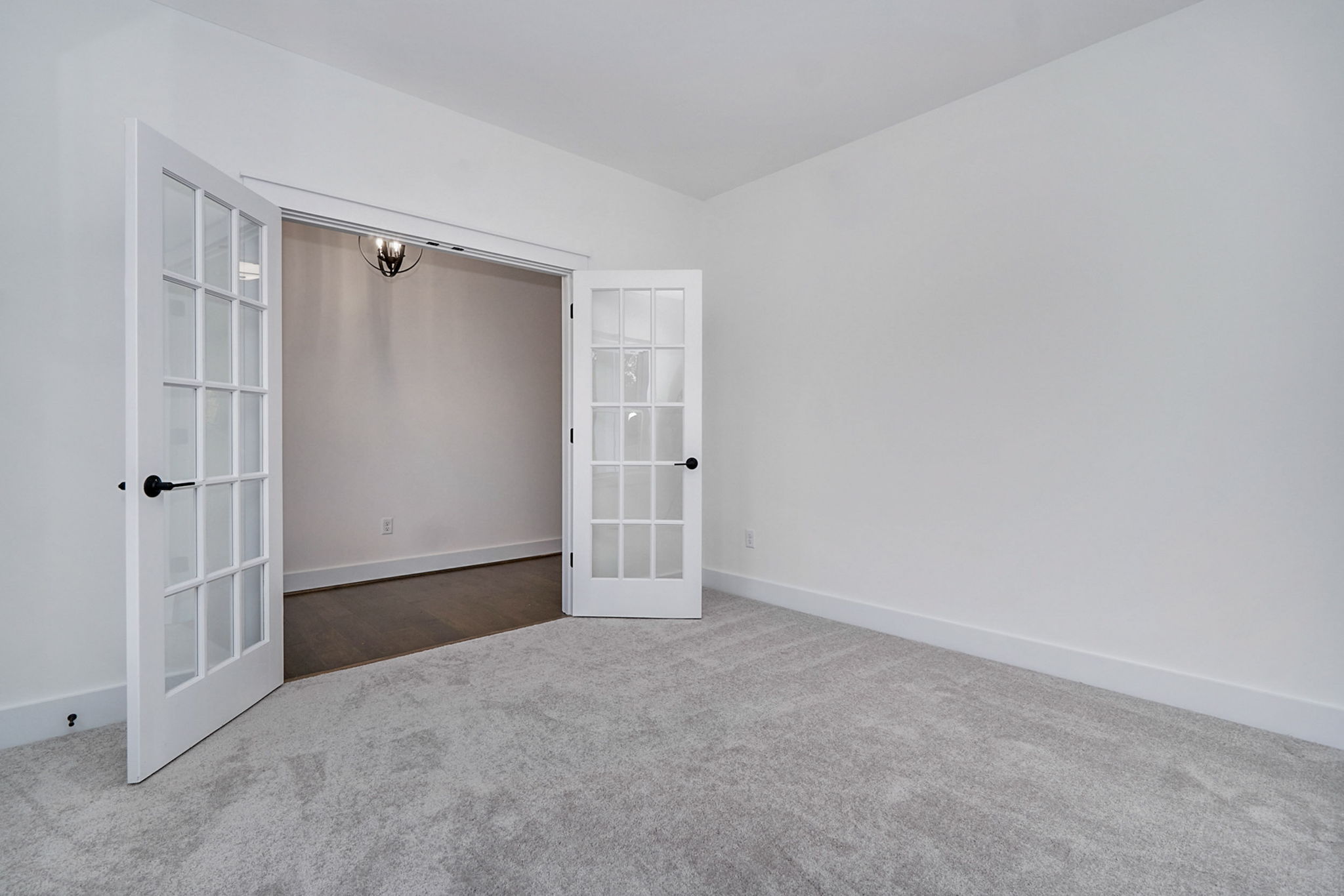 A room with light gray carpeting, white walls, and double glass French doors opening into another room with wooden flooring and a hanging light fixture.