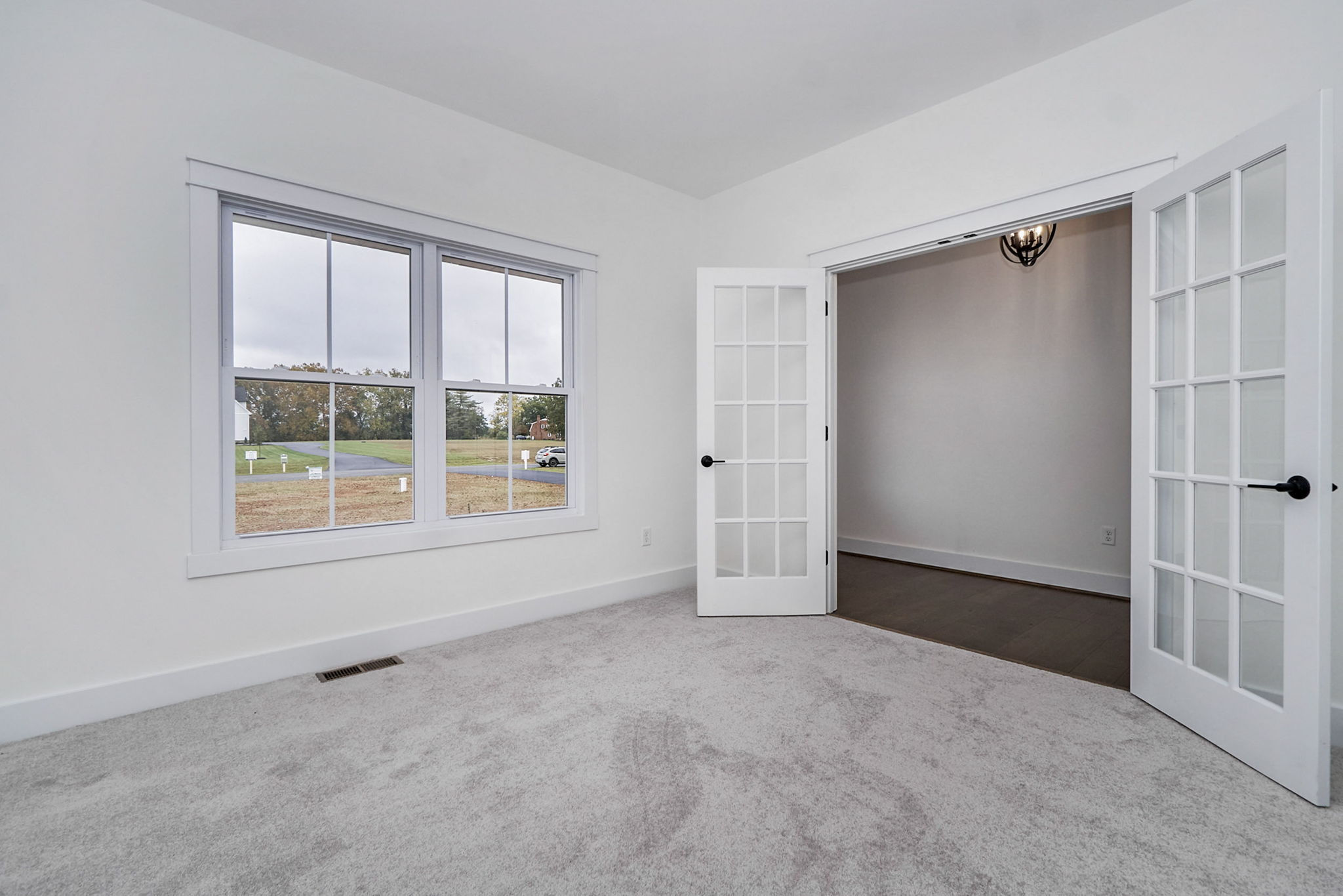 A bright, empty room with white walls and carpeted floor, featuring large windows and French doors leading to an adjacent space.