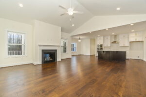 Spacious living room with hardwood floors, white walls, a fireplace, ceiling fan, and an adjoining kitchen area with dark cabinets.