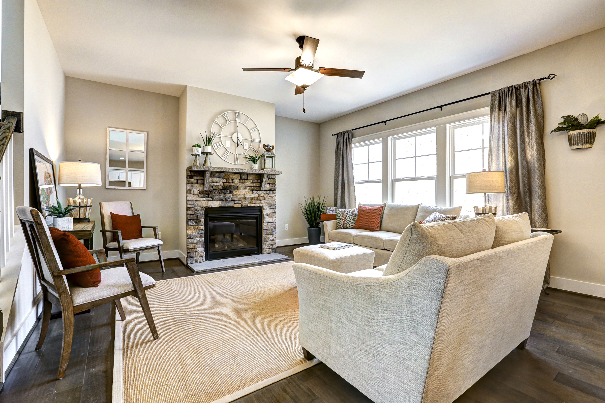 A well-lit living room features a large sofa, two armchairs, a fireplace with a stone surround, a wall-mounted clock, and large windows. The room is decorated with plants and neutral-colored decor.