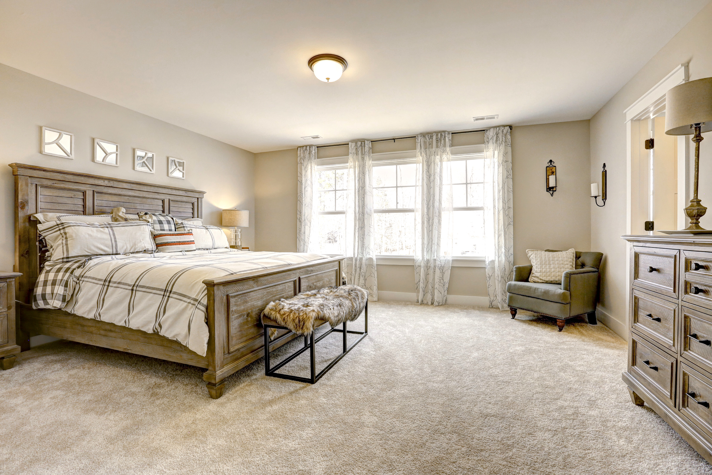 A cozy bedroom with a wooden bed, plaid bedding, fur bench, armchair, dresser, and large windows with sheer curtains. Walls are light beige with four framed decorations above the bed.