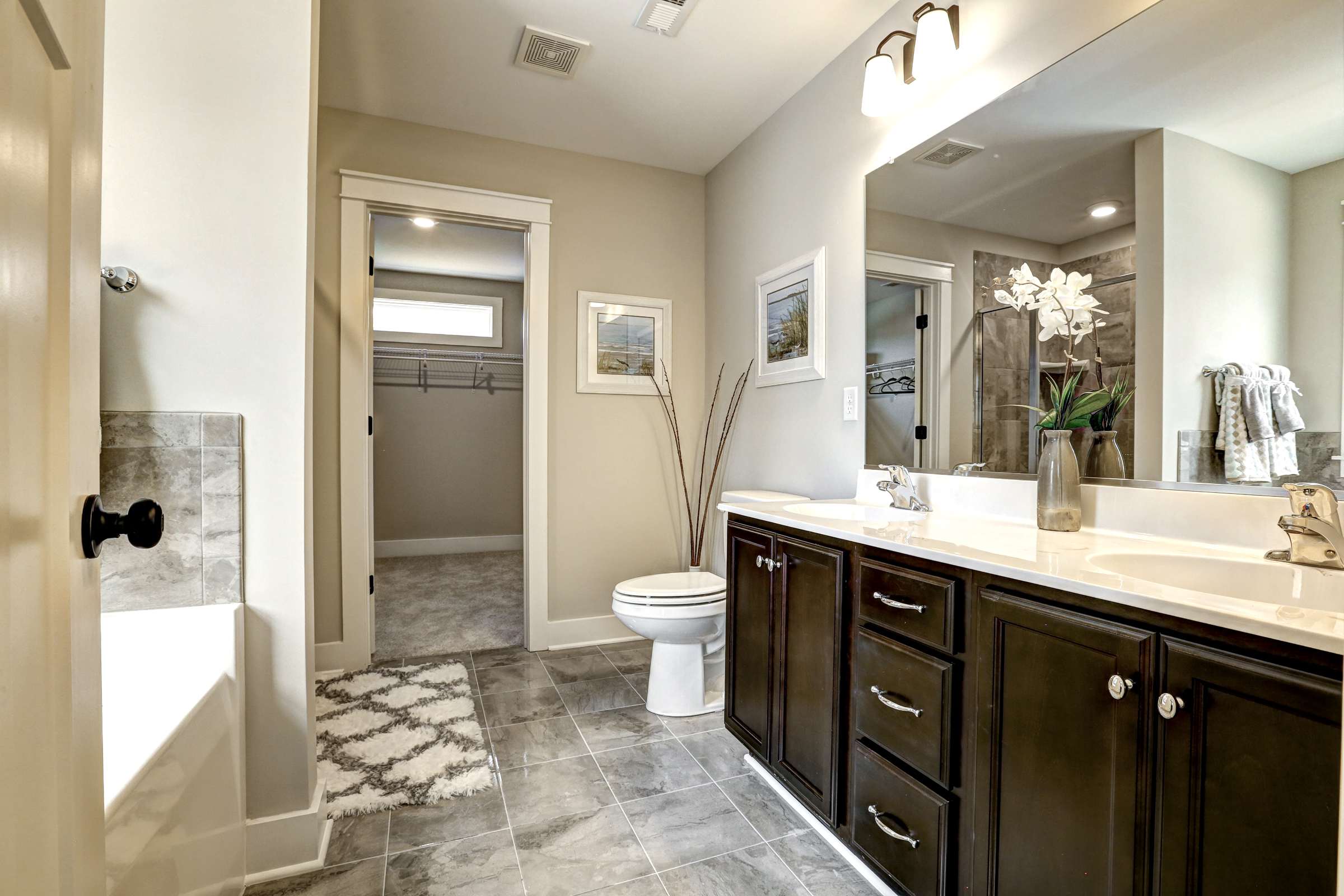 Modern bathroom with double sink vanity, large mirror, recessed lighting, bathtub, and toilet. There are framed pictures on the wall and a large closet is visible through an open door.