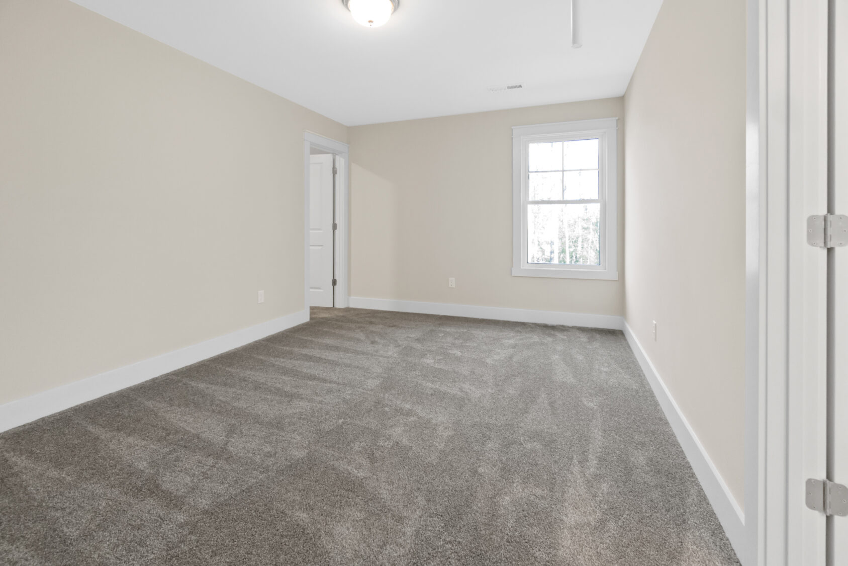 A vacant room with beige walls, white trim, a window, and gray carpet. A closed door is visible on one side.