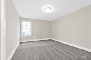 Empty room with gray carpet, light beige walls, and a ceiling light fixture. A single window with white trim allows natural light in.