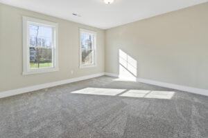 Empty room with beige walls, two windows, and gray carpet. Sunlight casts shadows on the carpet.