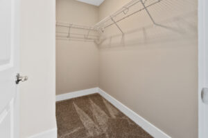 Empty walk-in closet with beige walls, white trim, and a gray carpeted floor. Two white wire shelves are installed on the left and back walls. Door is white with a silver handle.
