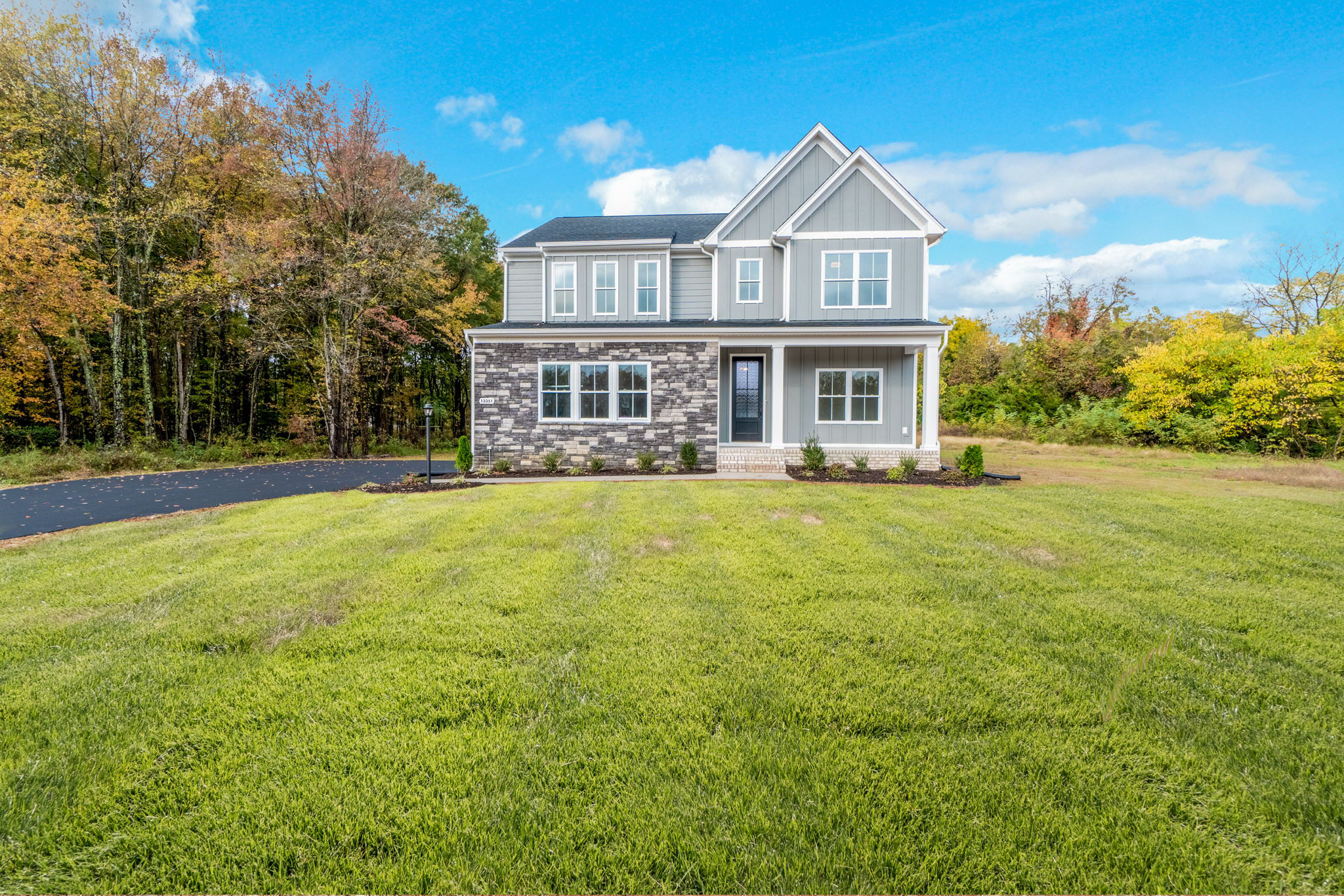 A modern two-story house with gray siding and a manicured lawn, situated next to a wooded area under a blue sky.