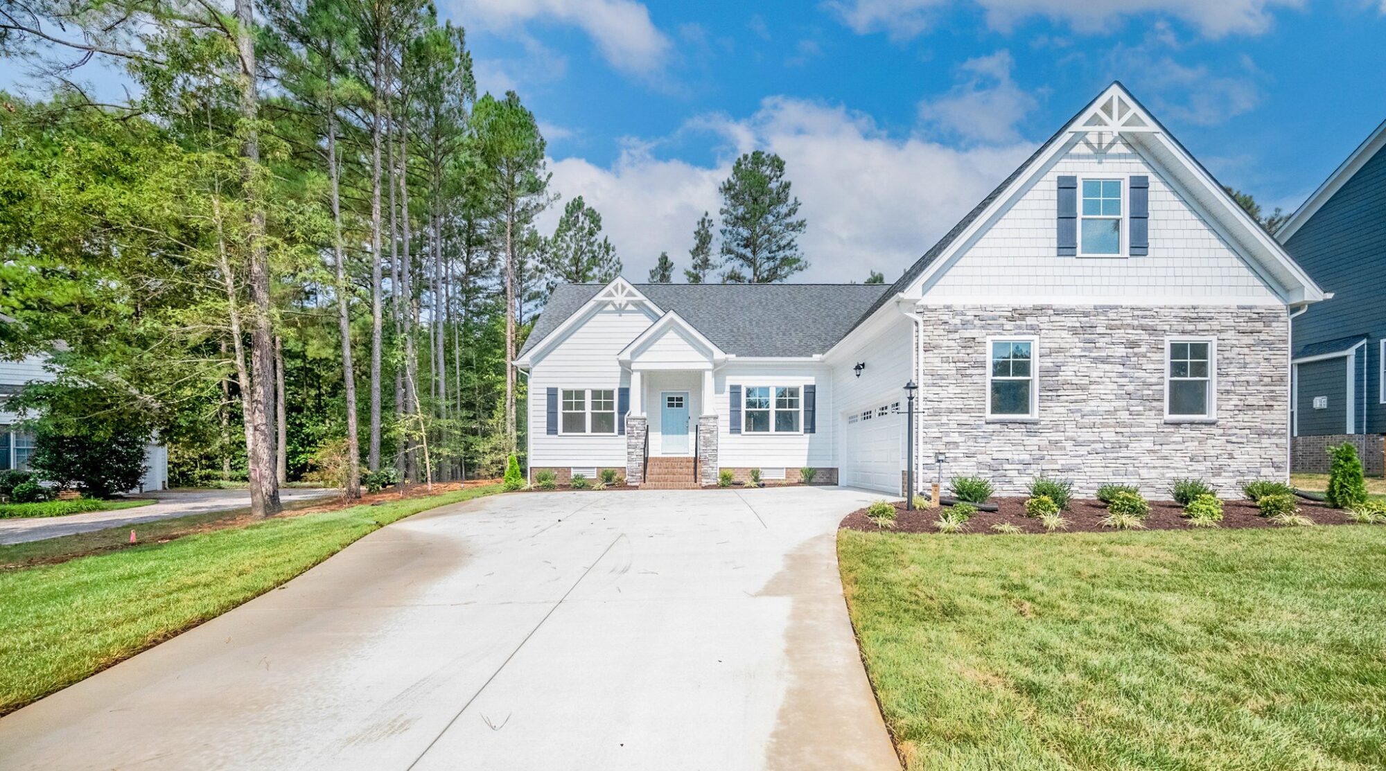 A two-story house with gray brick and white siding, a concrete driveway, a small front yard, and surrounded by tall trees under a blue sky.