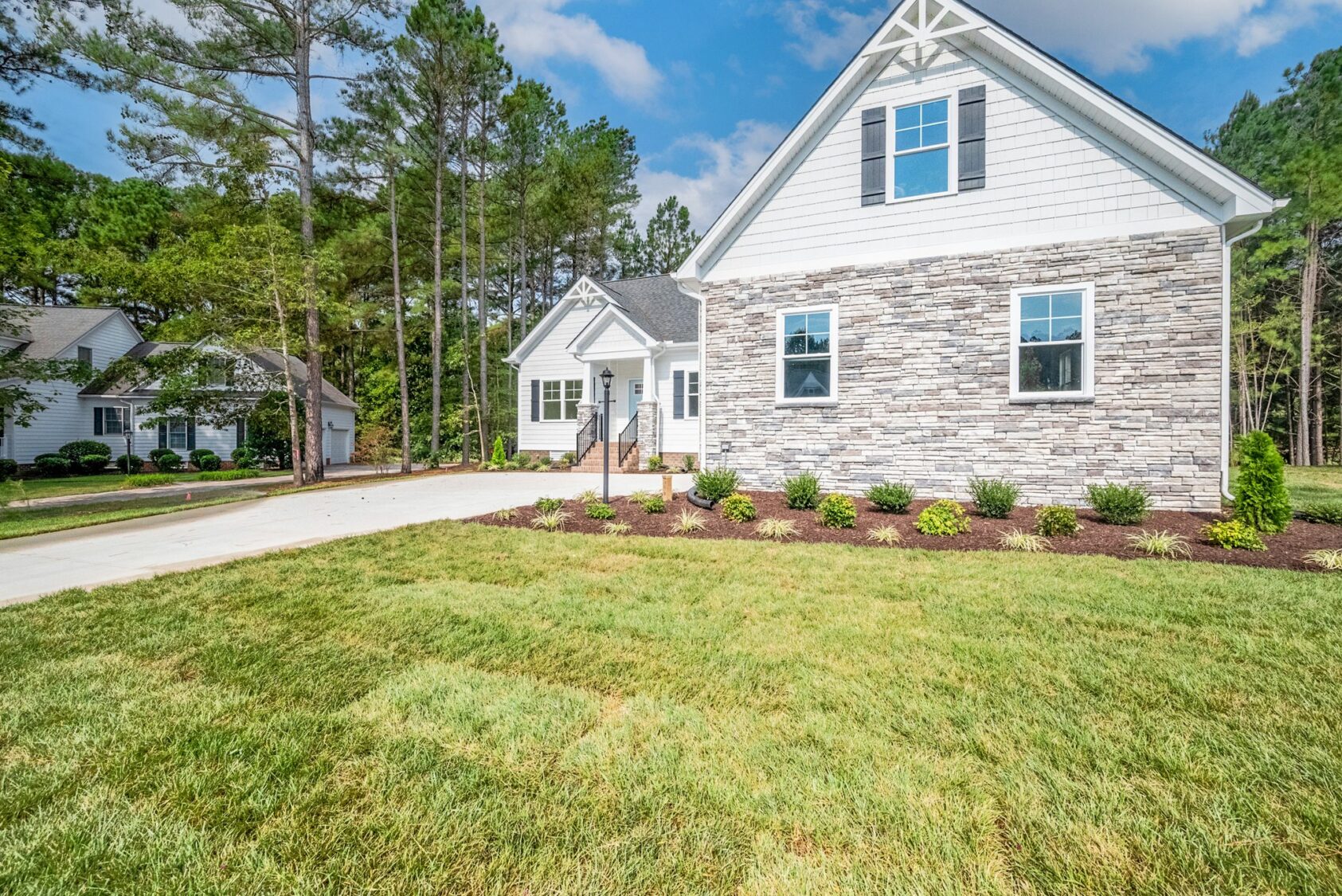A single-family home with stone and siding exterior, a concrete driveway, well-maintained lawn, and surrounding trees under a blue sky.