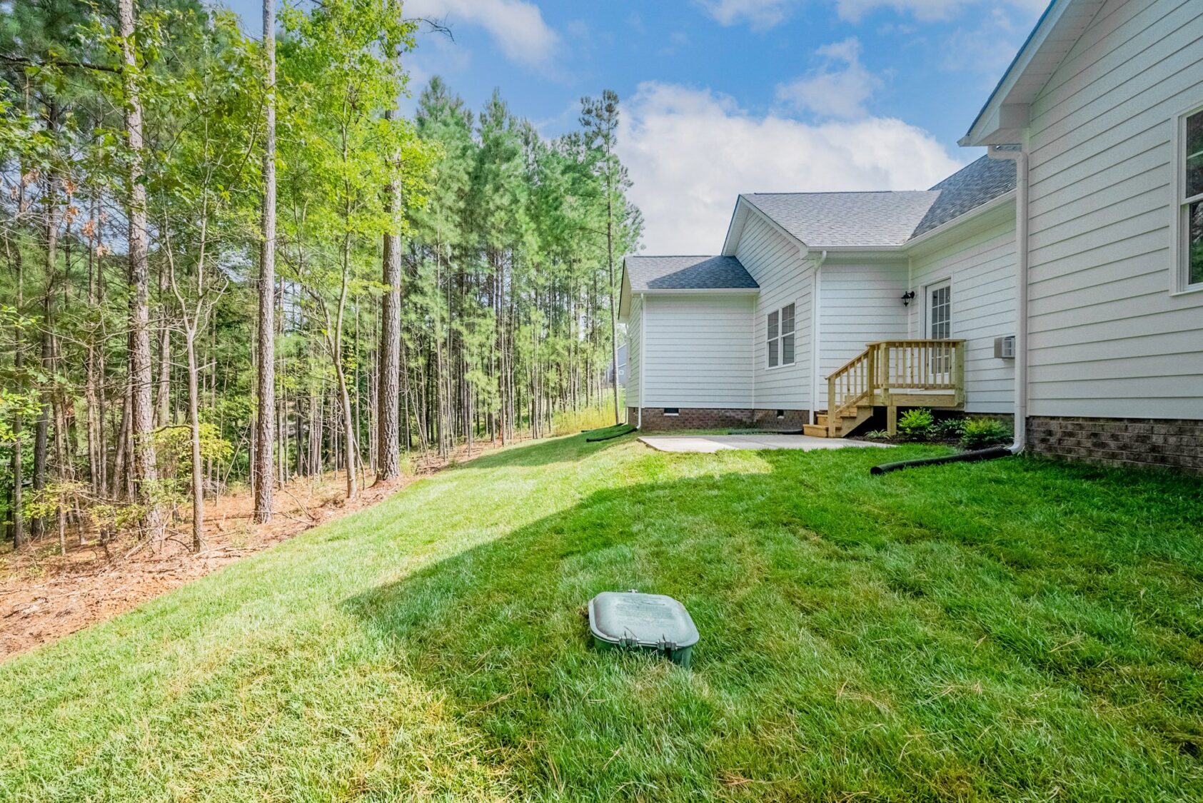 A grassy backyard of a white house with a small wooden deck, adjacent to a forested area.