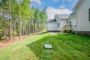 A grassy backyard of a white house with a small wooden deck, adjacent to a forested area.