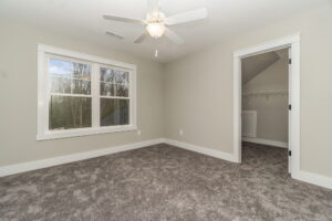 An empty room with beige walls and carpet, a ceiling fan, a window, and an open closet door.