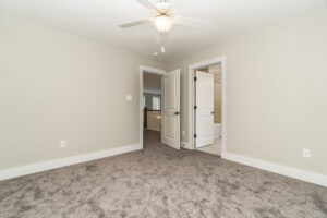 Empty room with beige walls, carpeted floor, ceiling fan, and two open doors leading to a hallway and bathroom.