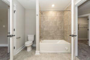 Bathroom with a bathtub and shower, beige tiled wall, toilet, and gray tile floor. White doors to the left and right lead to carpeted areas.