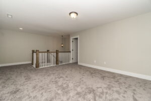 Empty carpeted room with light gray walls, a recessed light fixture, and a railing around a staircase opening. A closed door is on one wall.